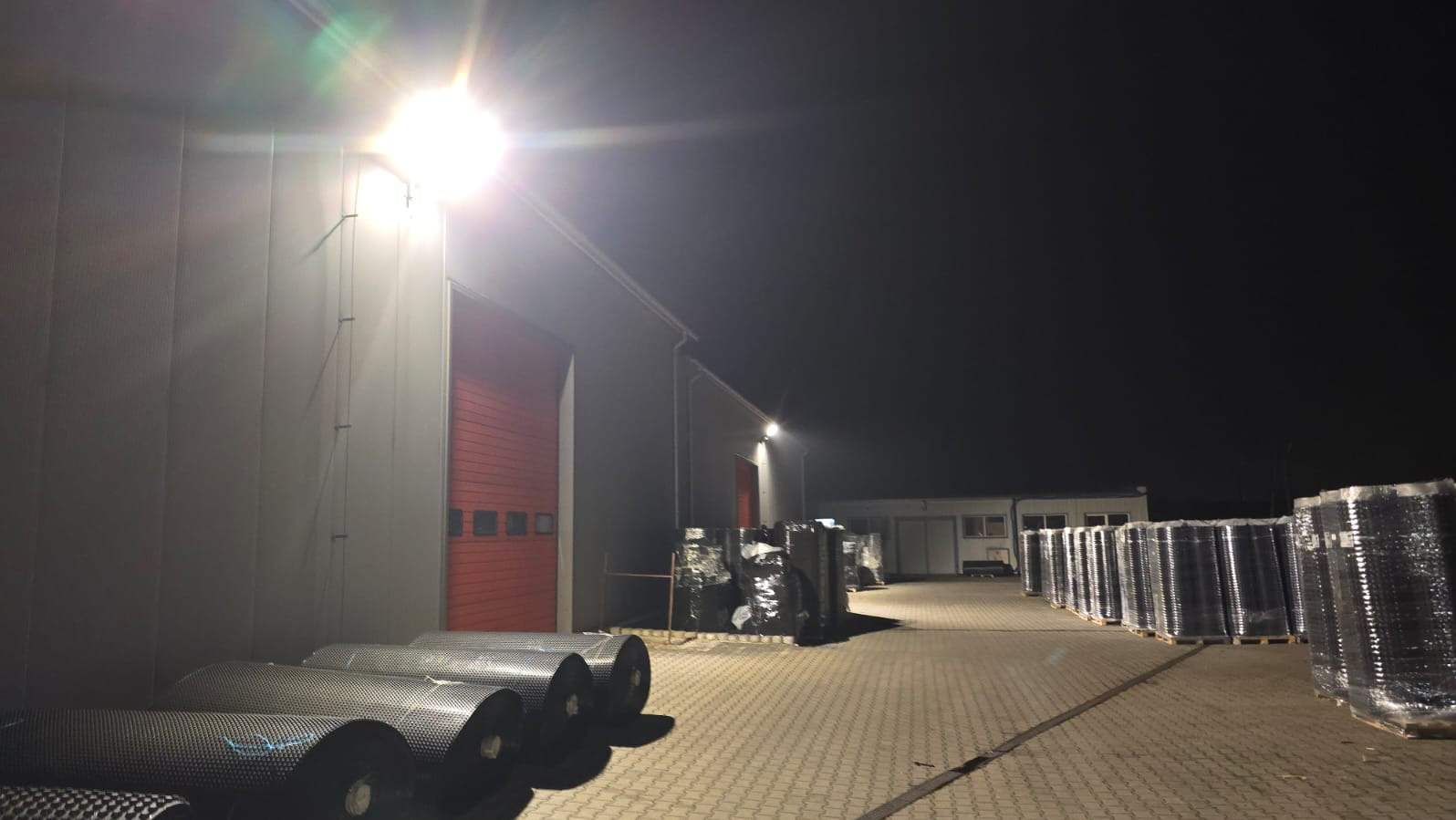 Nocne oświetlenie przemysłowe: jasna lampa halogenowa oświetla magazyn z czerwonymi bramami i paletami z materiałami budowlanymi na zewnątrz.