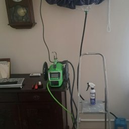 Klimatak Szymon Jabłoński - Czyszczenie klimatyzatora w domu: klimatyzator zabezpieczony folią ochronną z podpiętym urządzeniem do czyszczenia, drabina z butelką środka czyszczącego obok, na komodzie zegar i narzędzia.