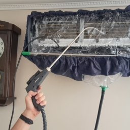 Klimatak Szymon Jabłoński - Czyszczenie klimatyzatora typu split za pomocą specjalistycznego urządzenia pod ciśnieniem, z założonym pokrowcem ochronnym zbierającym brud, obok wisi stary zegar.