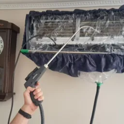 Czyszczenie klimatyzatora typu split za pomocą specjalistycznego urządzenia pod ciśnieniem, z założonym pokrowcem ochronnym zbierającym brud, obok wisi stary zegar.