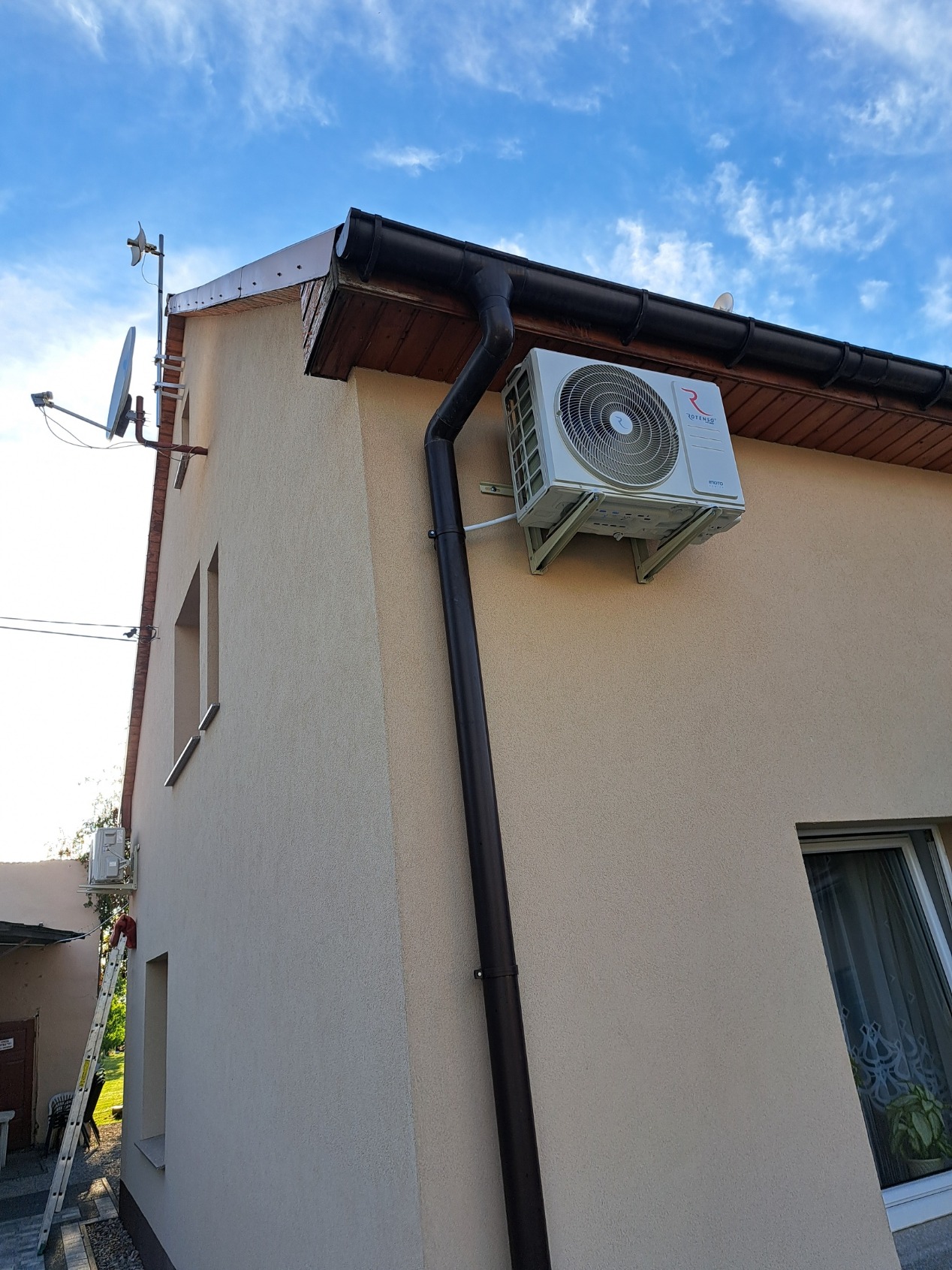 Biała jednostka zewnętrzna klimatyzatora Rotenso zamontowana na elewacji beżowego domu jednorodzinnego, widoczna rynna spustowa, antena satelitarna i drabina oparta o ścianę w tle.