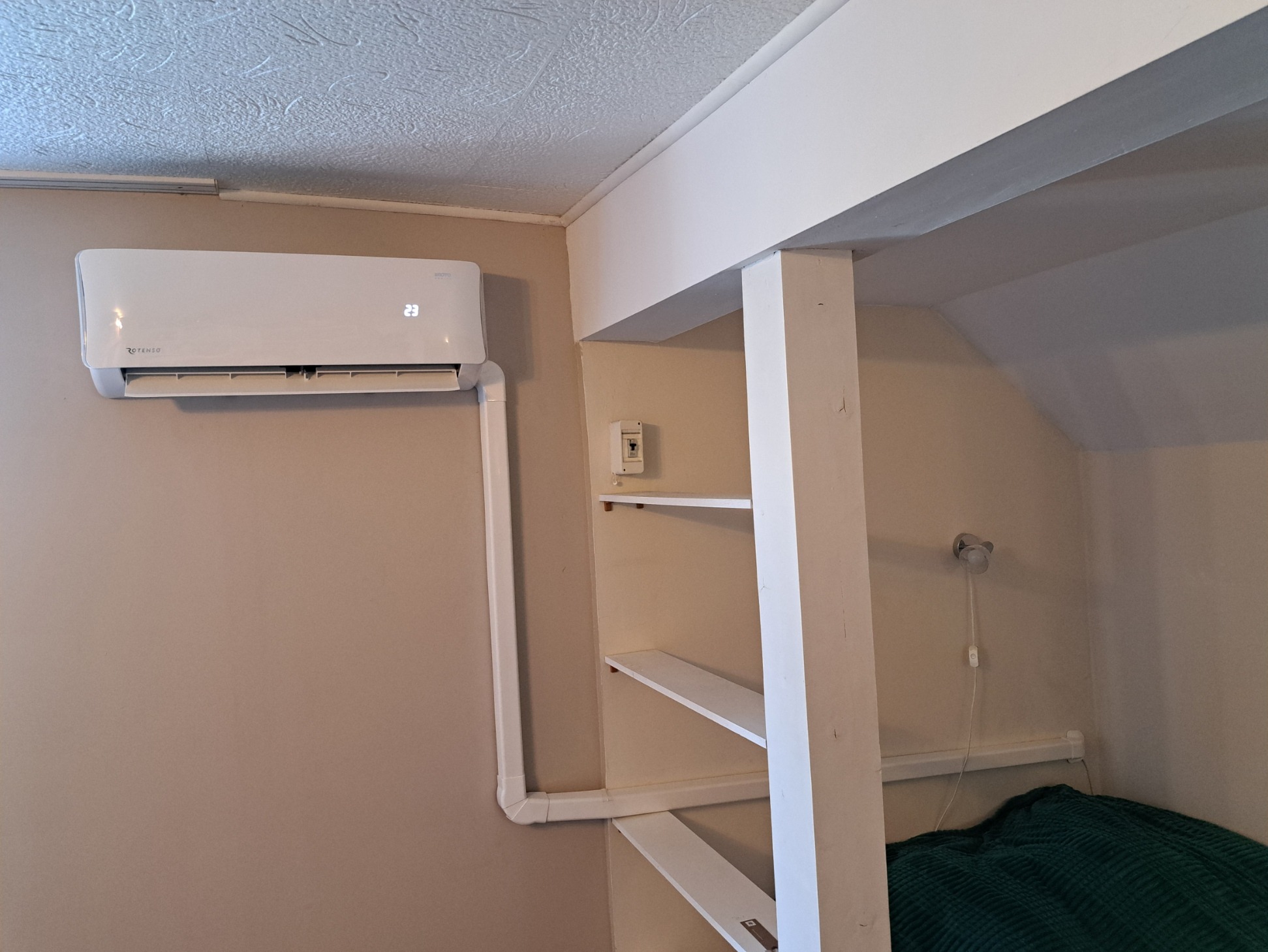 Biały klimatyzator Rotenso, wyświetlający 23 stopnie Celsjusza, zamontowany na beżowej ścianie w pokoju z antresolą, widoczne białe rury instalacyjne.