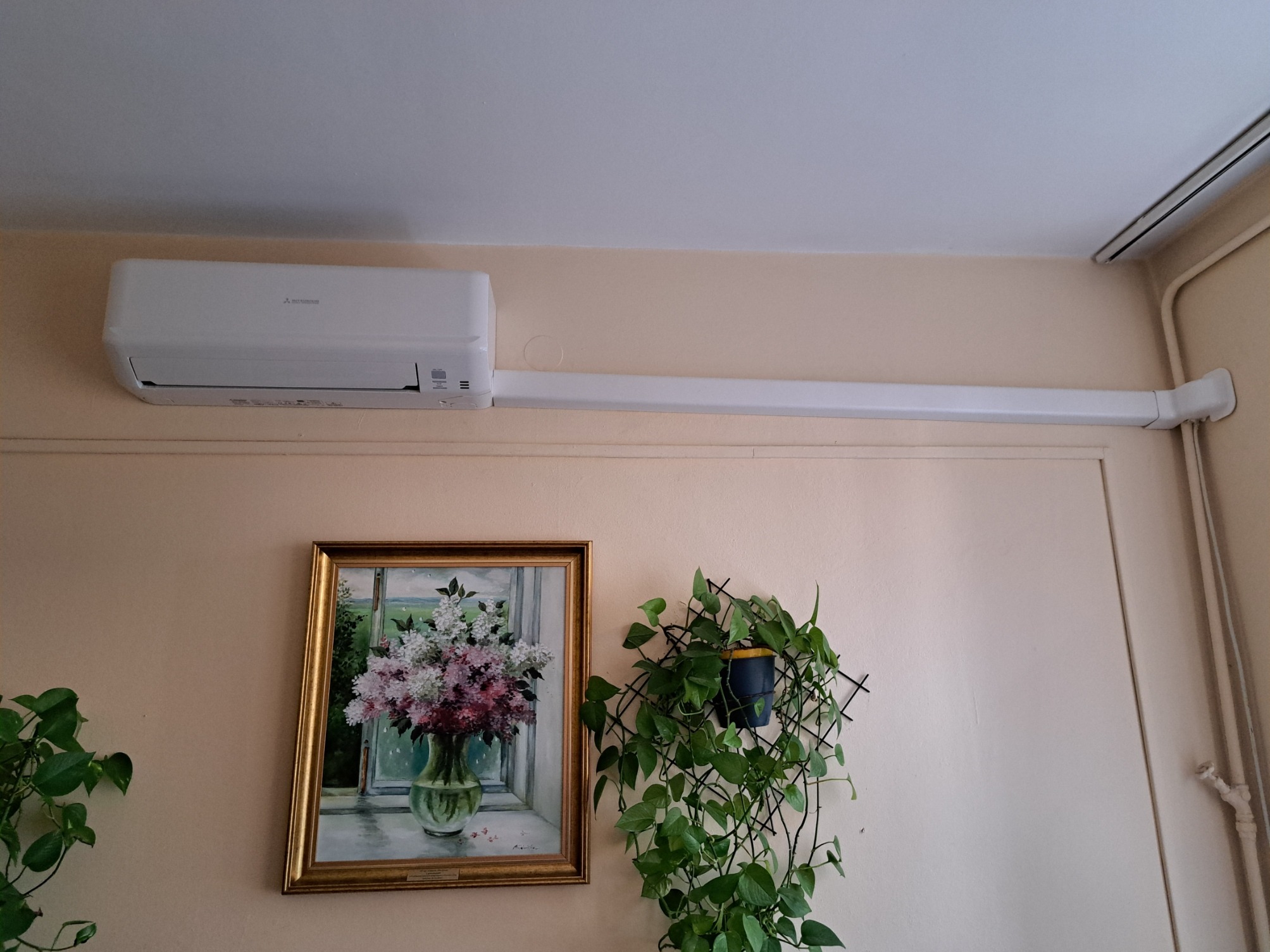 Biały klimatyzator Mitsubishi zamontowany na ścianie w pomieszczeniu, z widoczną instalacją prowadzącą wzdłuż ściany, obok wiszący obraz z kwiatami w złotej ramie i zielona roślina doniczkowa...