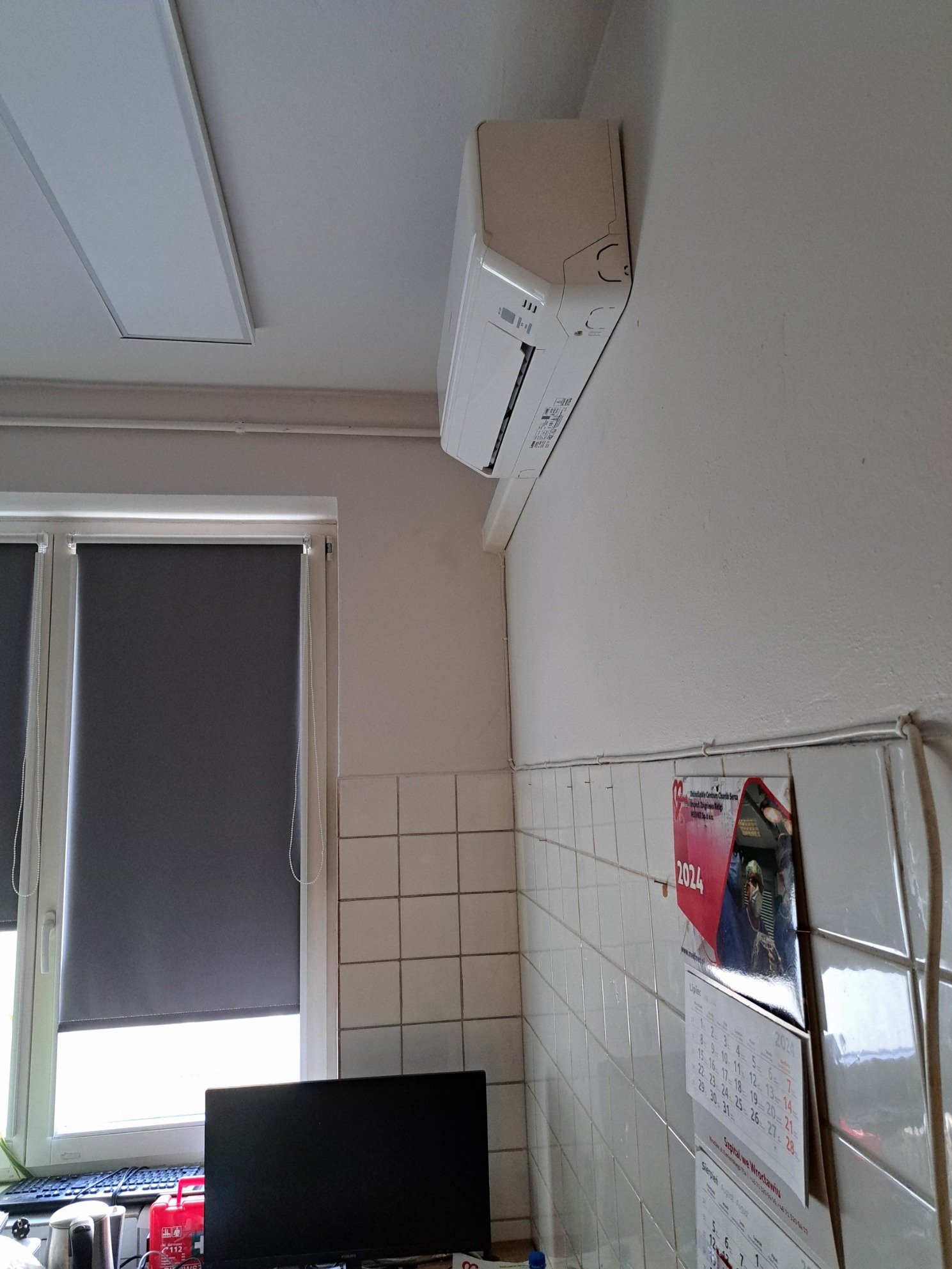 Biały klimatyzator ścienny zamontowany w pomieszczeniu z kafelkową ścianą, widoczne okno z roletą i kalendarz na ścianie.