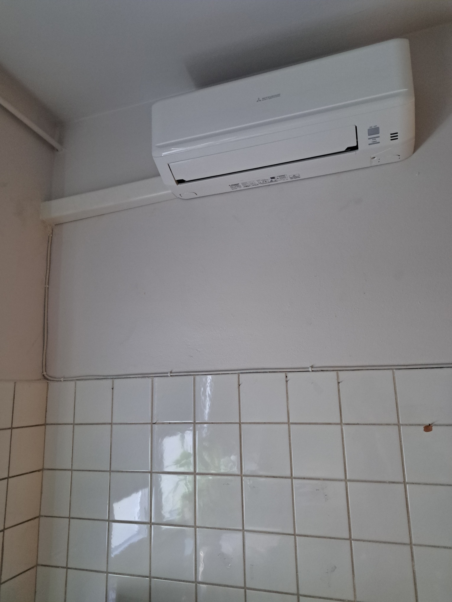 Biały klimatyzator Mitsubishi zamontowany na ścianie nad kafelkami w jasnym pomieszczeniu. Widoczne białe rury instalacyjne biegnące wzdłuż ściany.