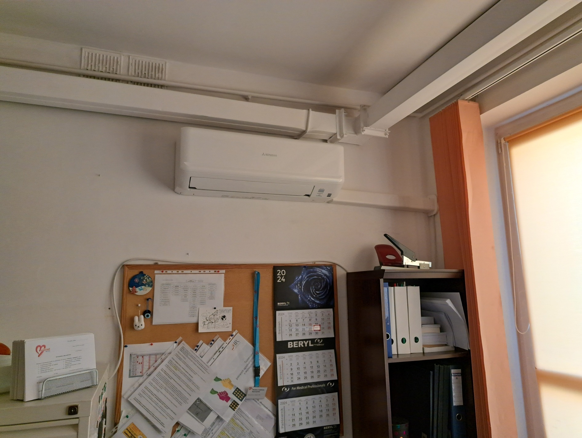 Biały klimatyzator Mitsubishi Electric zamontowany na ścianie nad biurkiem z kalendarzem Beryl w pomieszczeniu biurowym.