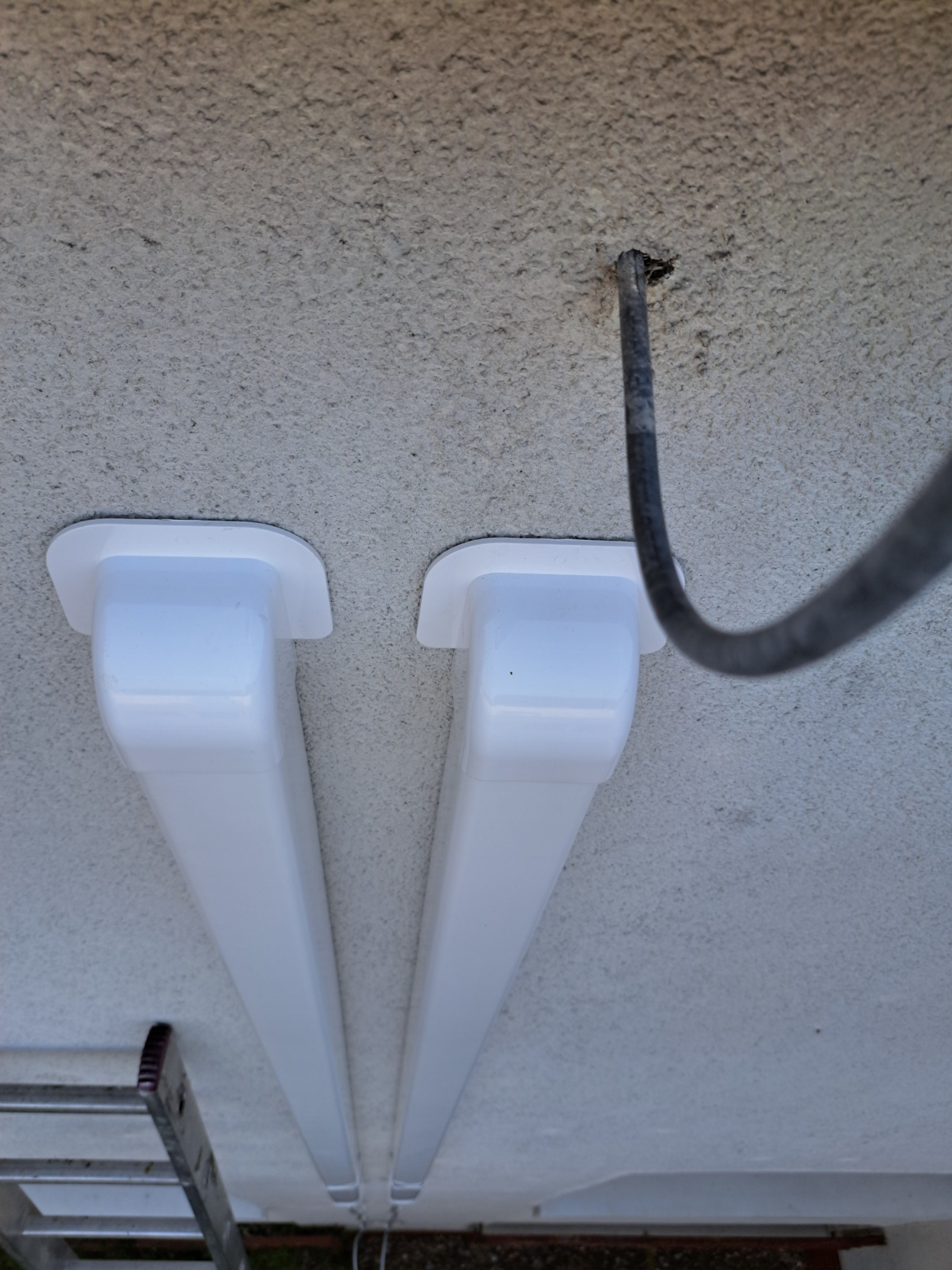 Dwa białe kanały wentylacyjne przymocowane do szorstkiego sufitu, widoczny kabel i fragment drabiny.