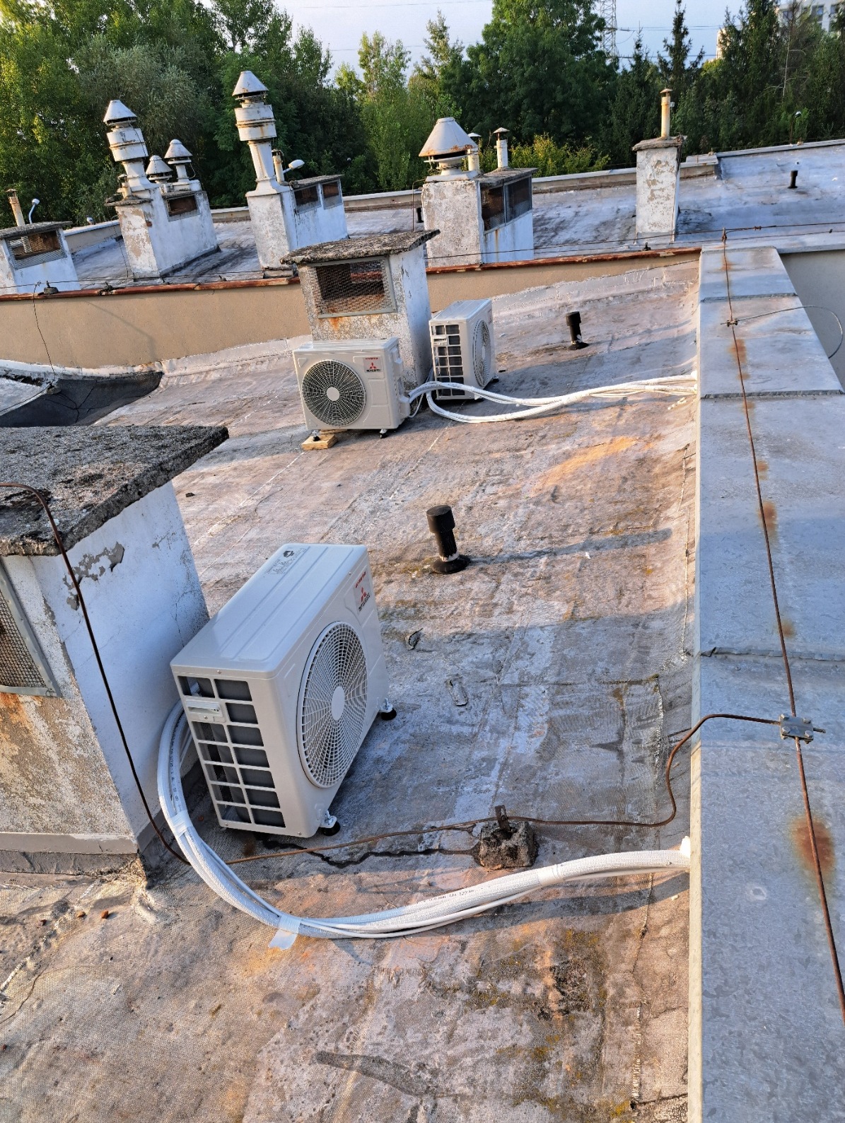 Trzy jednostki zewnętrzne klimatyzacji Mitsubishi na płaskim, betonowym dachu budynku z kominami, widoczne białe rury instalacyjne.