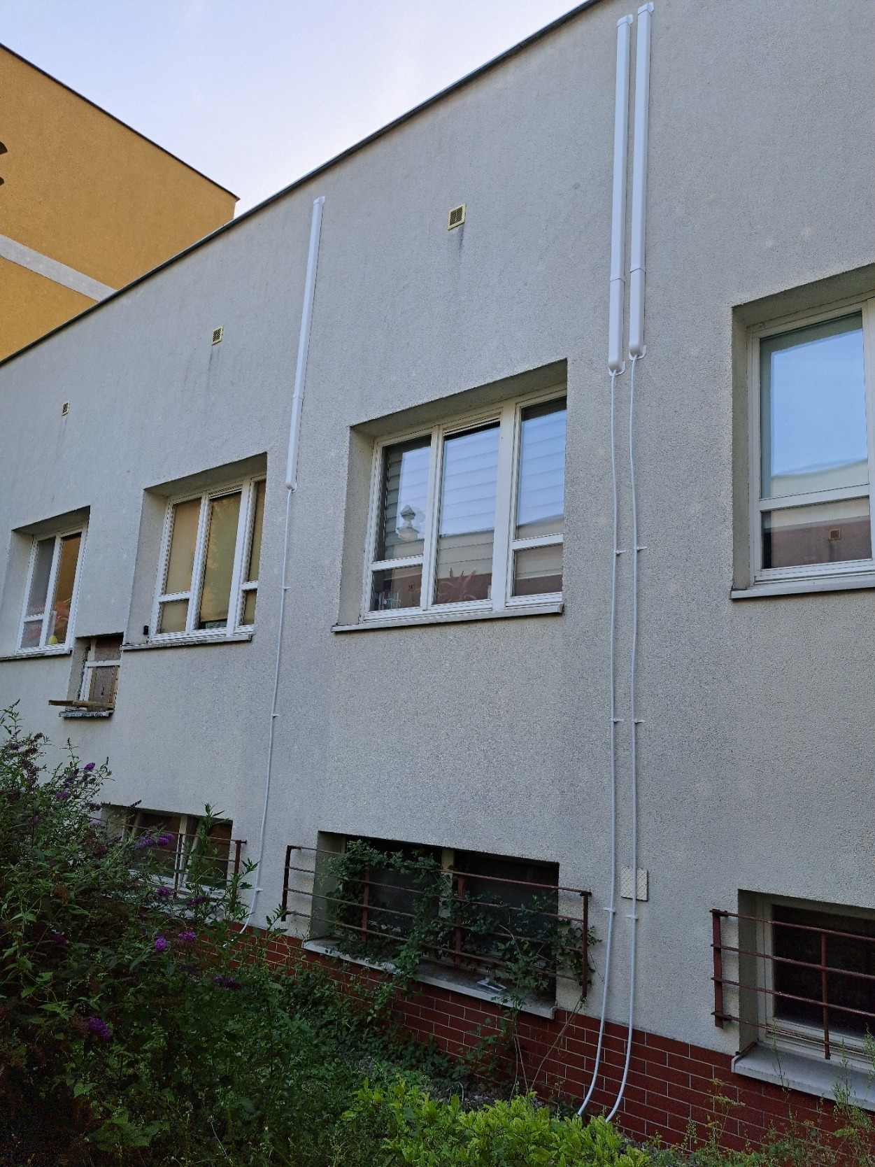 Szary budynek z oknami, z widocznymi białymi rurami klimatyzacji na elewacji, z zielenią przed oknami na parterze.