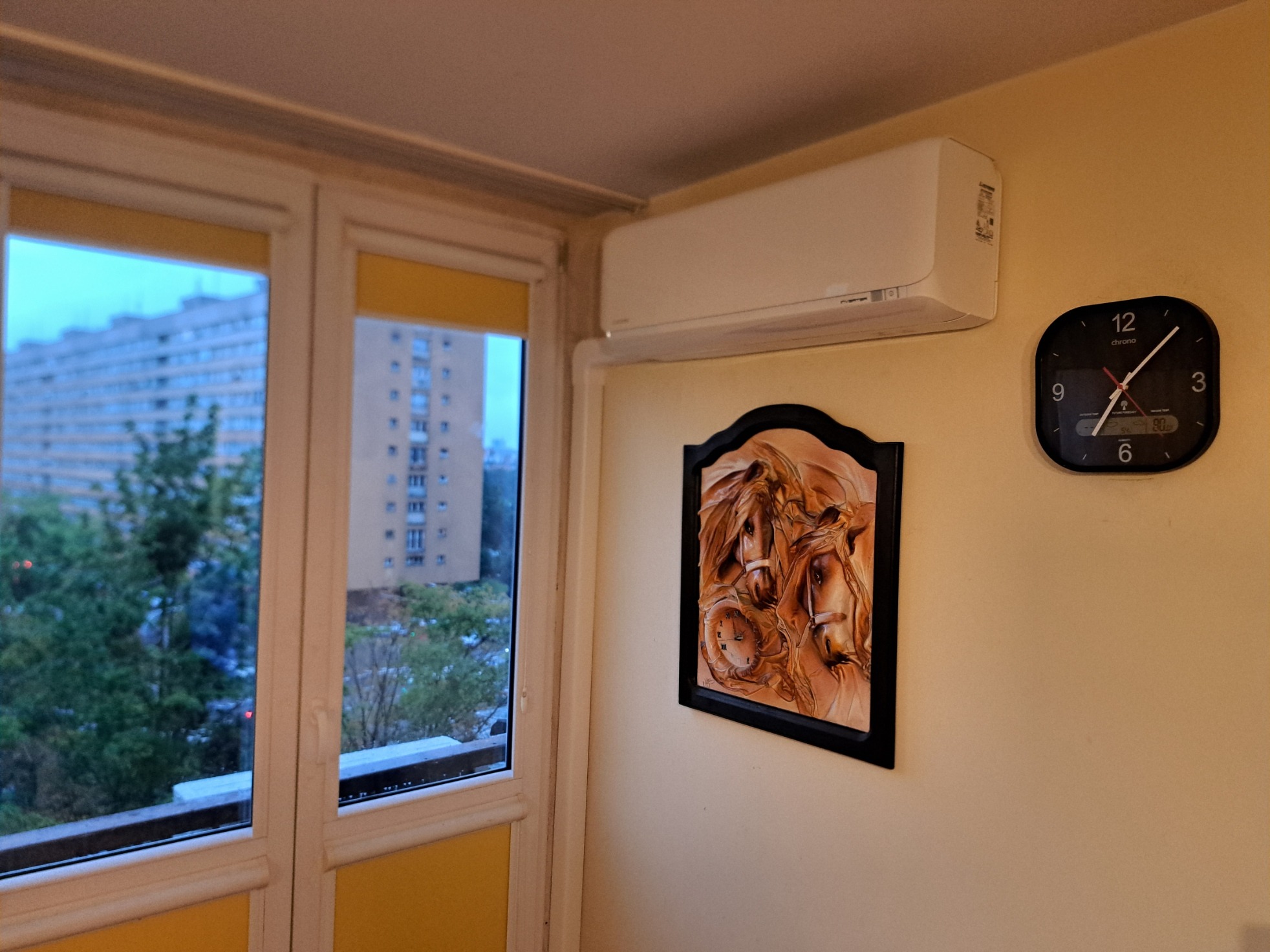 Biała jednostka klimatyzacyjna na ścianie w mieszkaniu, obok wiszący zegar i obraz z motywem koni, widok z okna na blok.