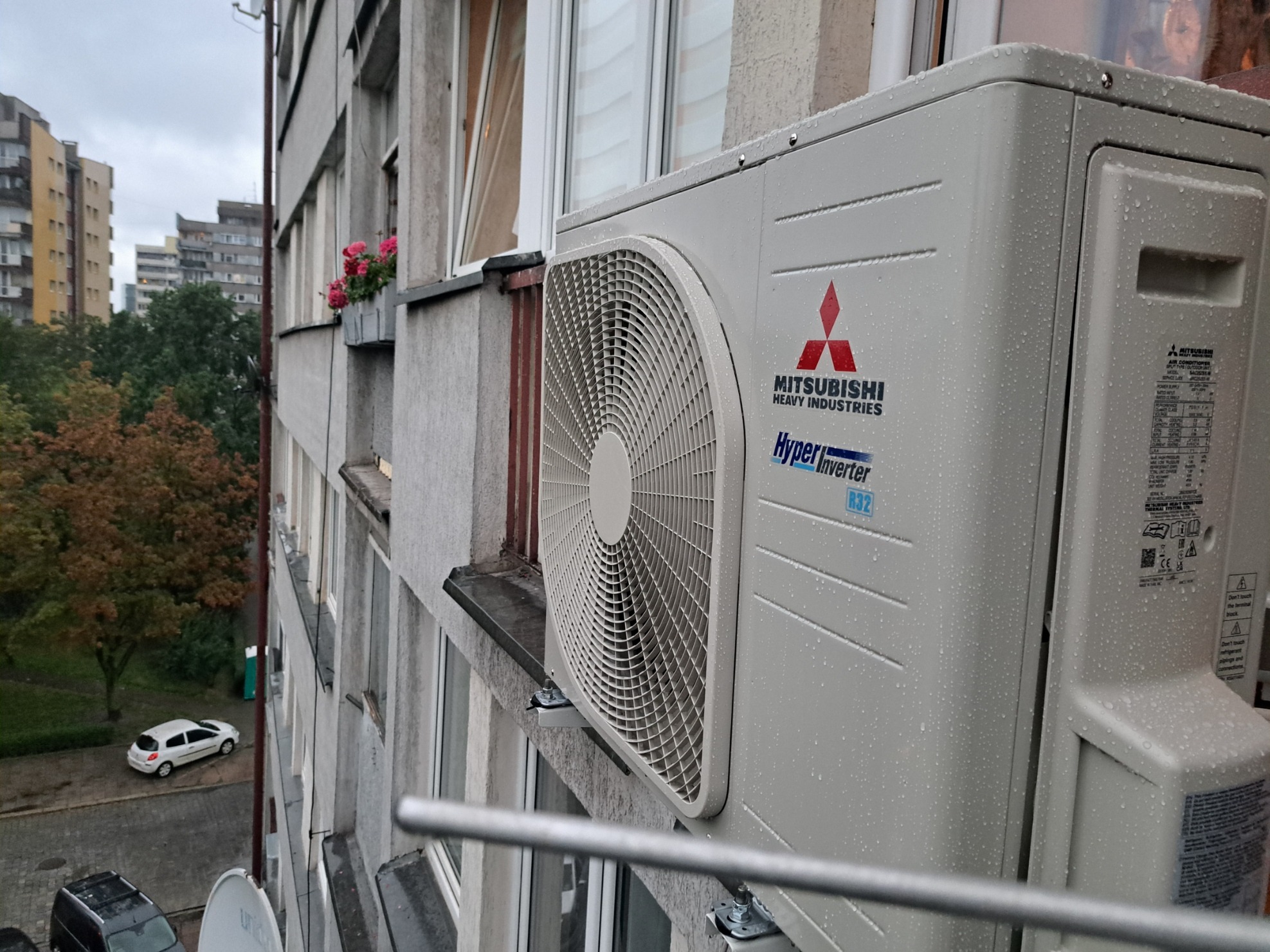 Zewnętrzna jednostka klimatyzacji Mitsubishi Heavy Industries zamontowana na ścianie bloku mieszkalnego, widok z balkonu na osiedle w pochmurny dzień.