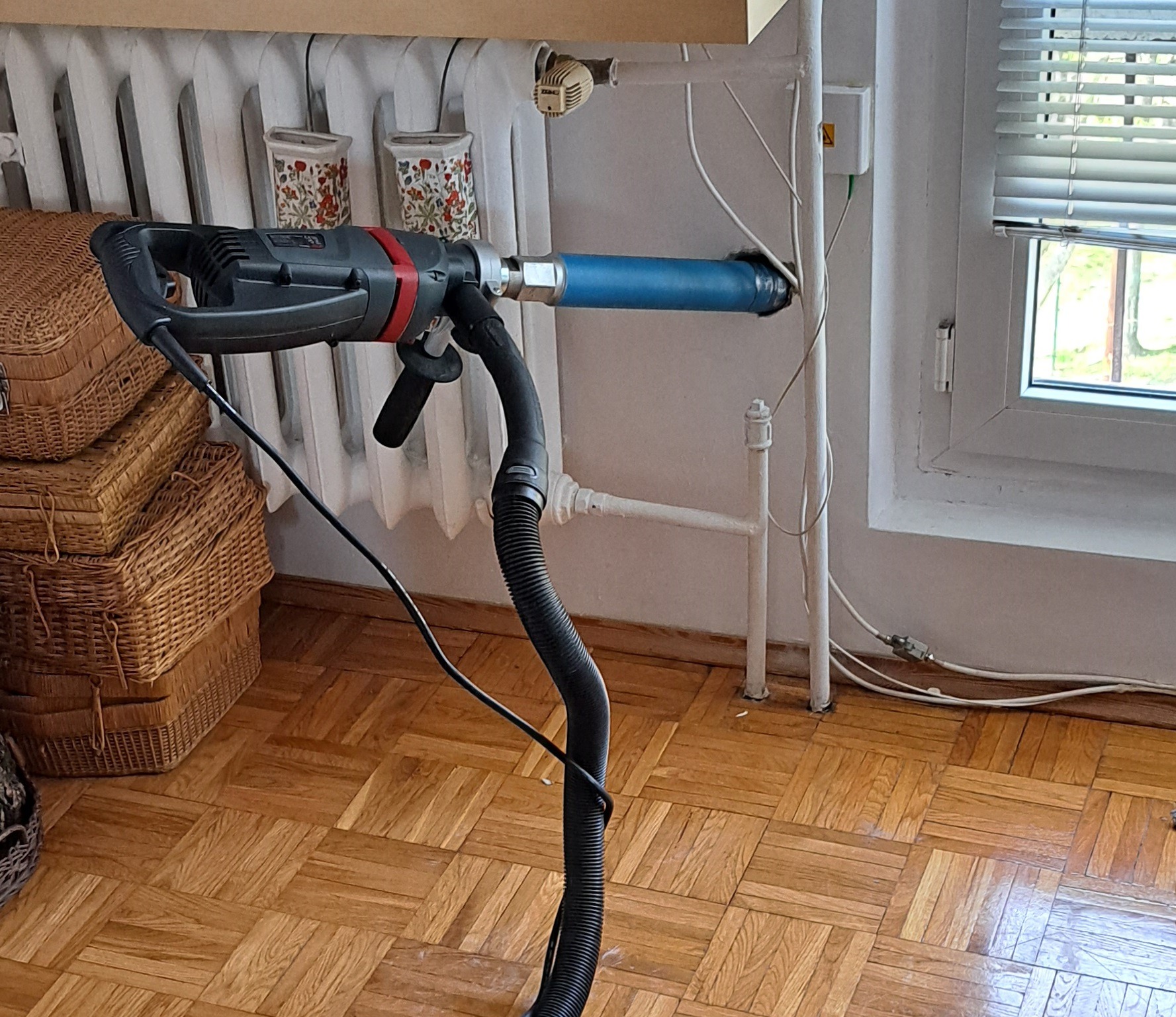 Wiertarka z odkurzaczem podłączona do otworu w ścianie obok grzejnika i okna z żaluzjami, zbierająca pył podczas wiercenia.