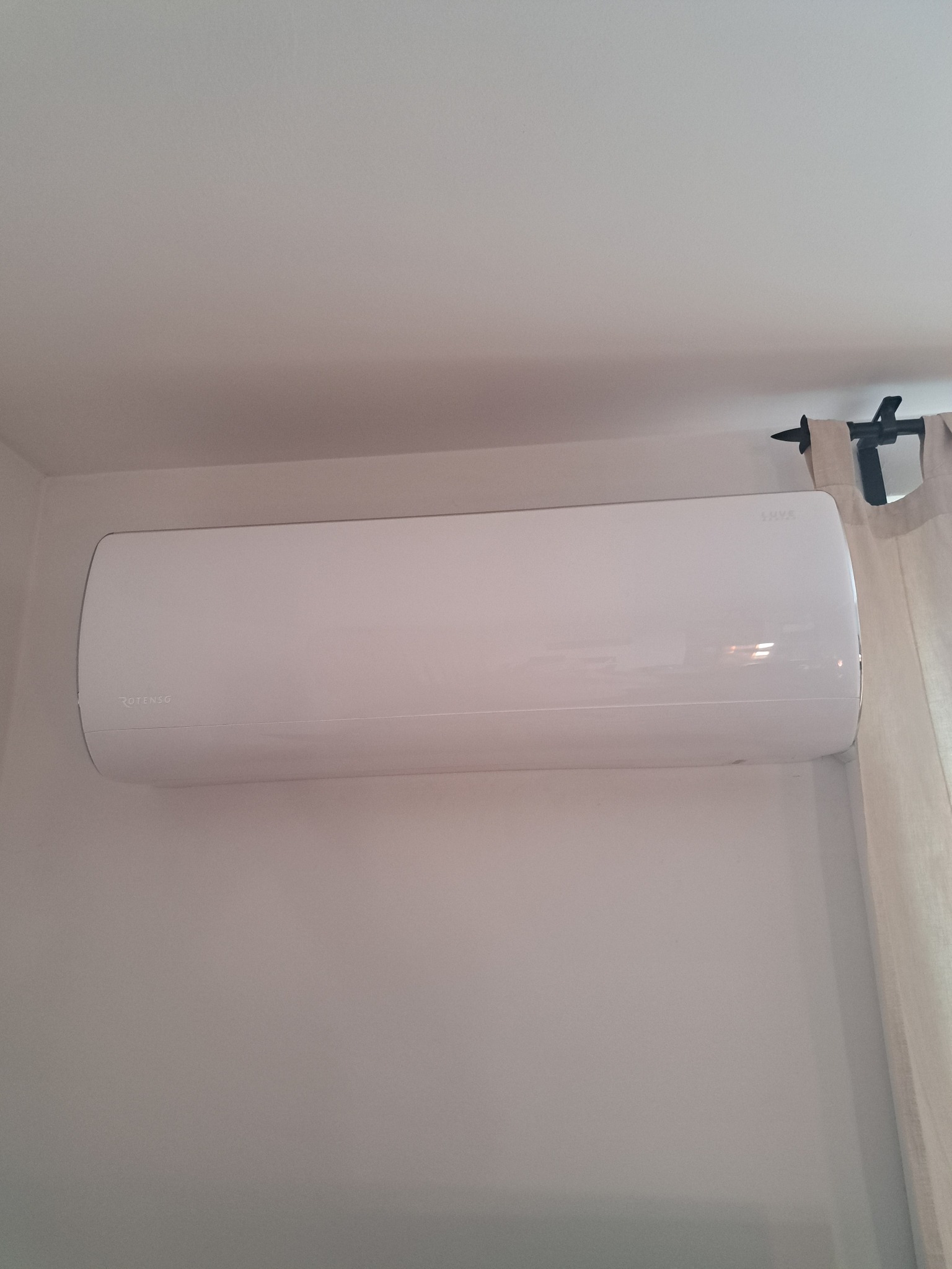 Biały klimatyzator marki Rotenso, zamontowany na białej ścianie w pomieszczeniu z zasłoną w tle.