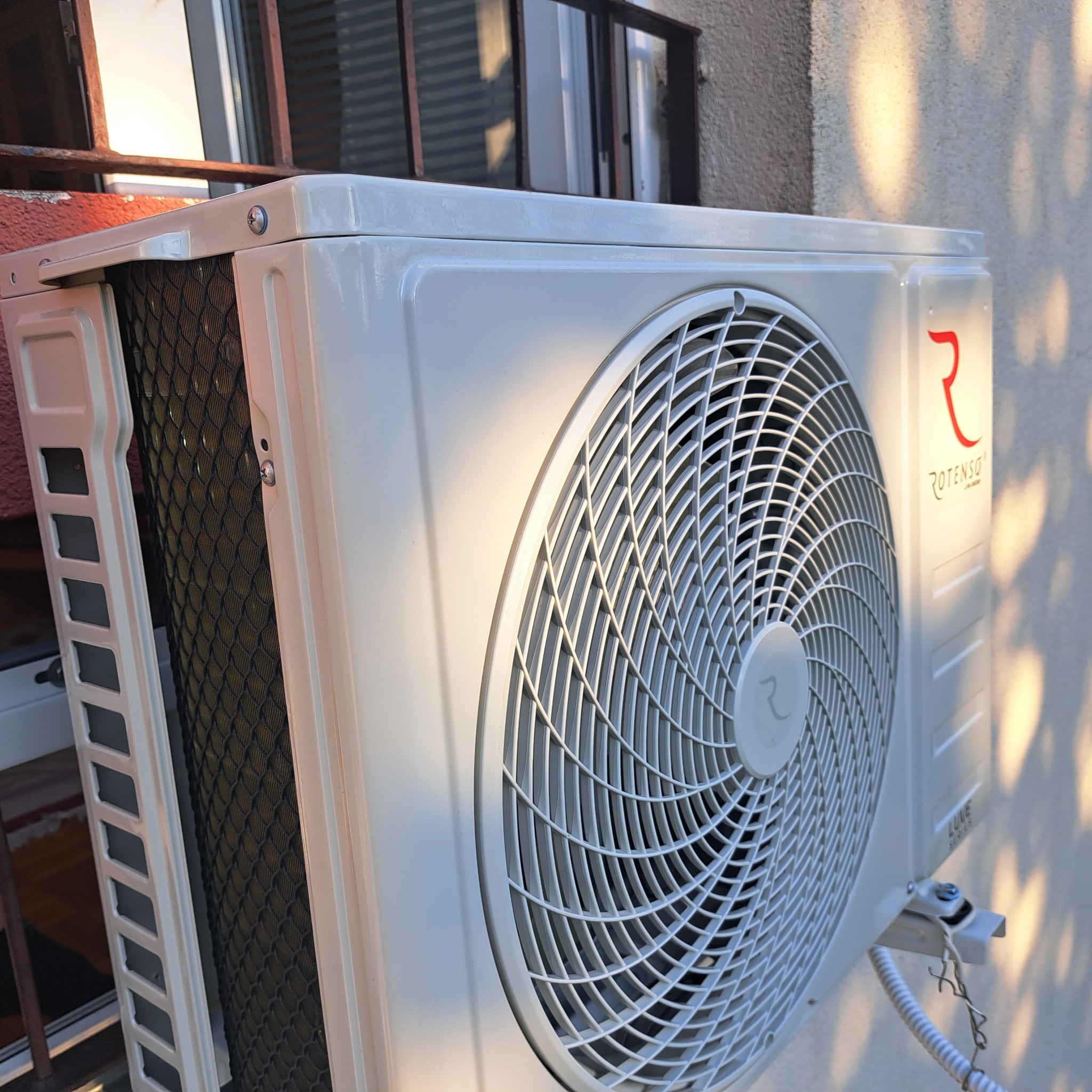 Zewnętrzna jednostka klimatyzacji Rotenso Luxe Series zamontowana na ścianie budynku, widoczne cieniowanie drzew na elewacji.