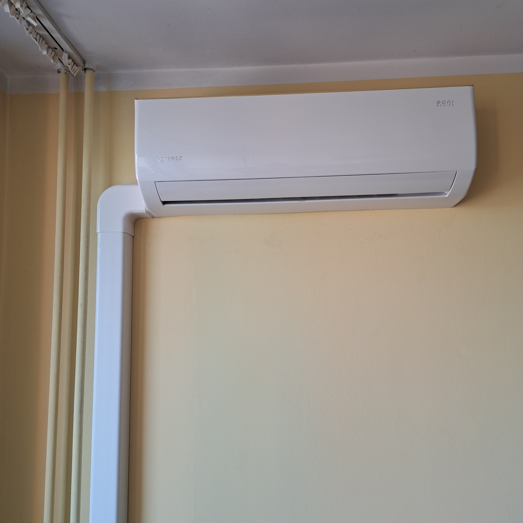 Biały klimatyzator marki Rotenso Roni Series zamontowany na żółtej ścianie z widocznymi rurami odprowadzającymi skropliny w białej osłonie.