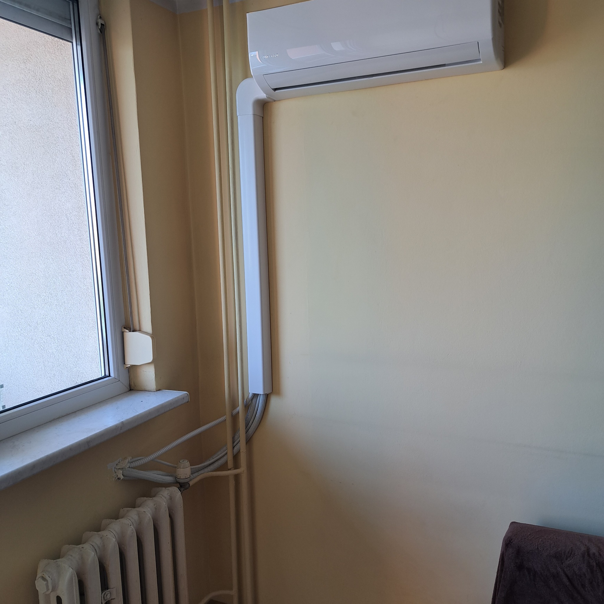 Biały klimatyzator ścienny zamontowany w pokoju, widoczne rury odprowadzające skropliny, obok okno i grzejnik.