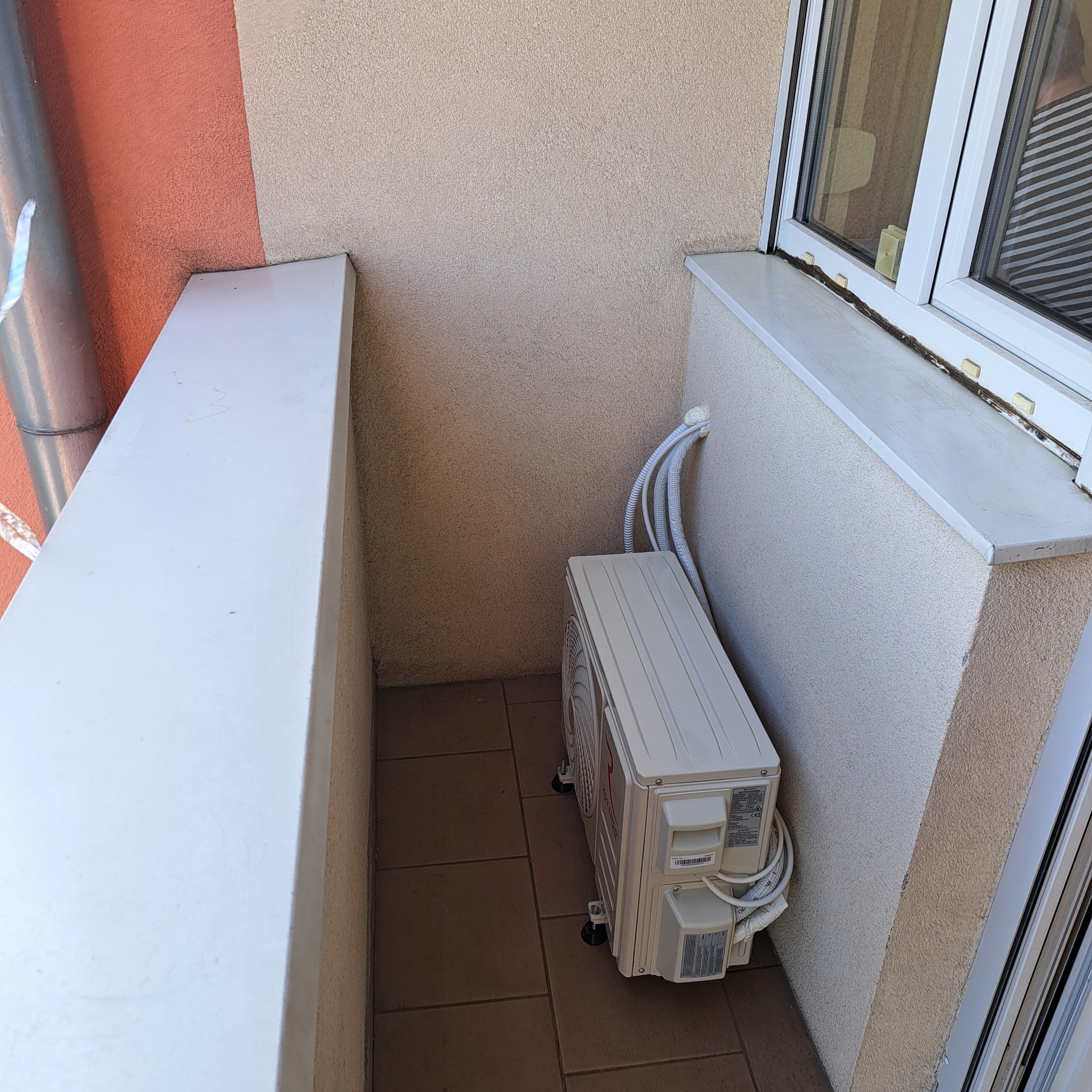 Zewnętrzna jednostka klimatyzacji zamontowana na balkonie, widoczne przewody wchodzące do budynku, brązowe płytki podłogowe i fragment okna.