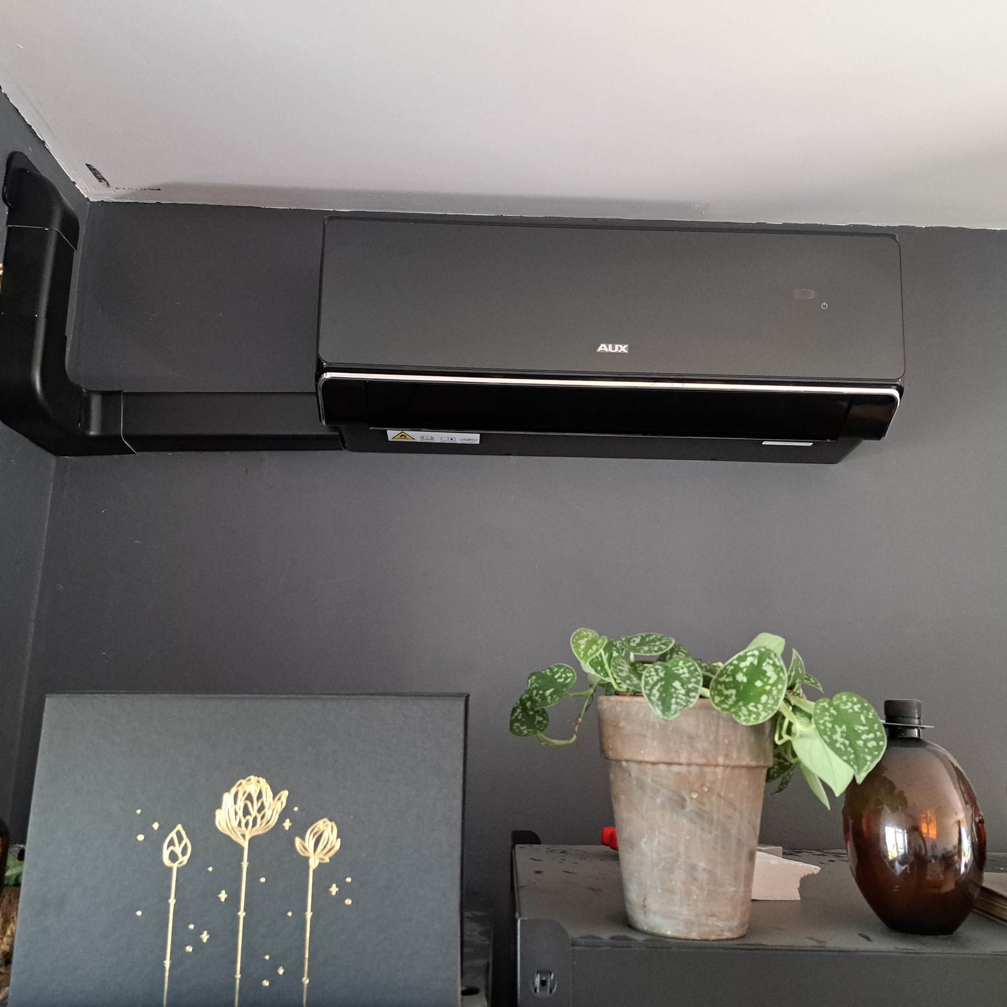 Czarna jednostka klimatyzacji AUX zamontowana na szarej ścianie w pomieszczeniu, poniżej dekoracje: roślina w doniczce, obrazek z motywem kwiatowym i brązowa butelka.