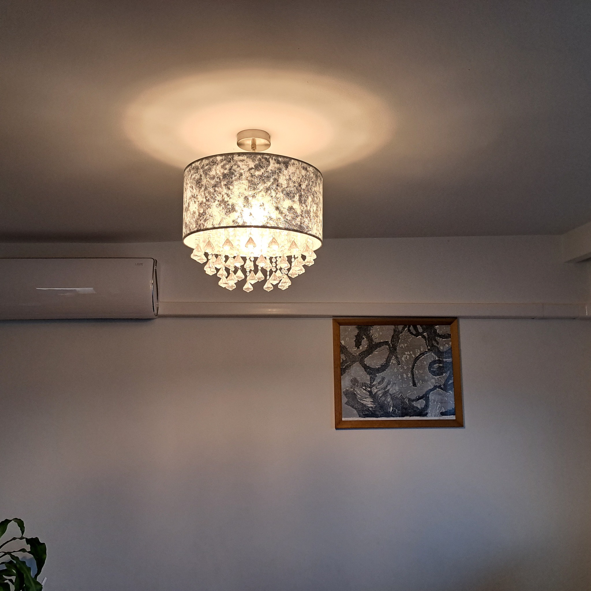 Biała ściana z zamontowanym klimatyzatorem, podwieszonym żyrandolem z kryształkami i obrazem w drewnianej ramie.