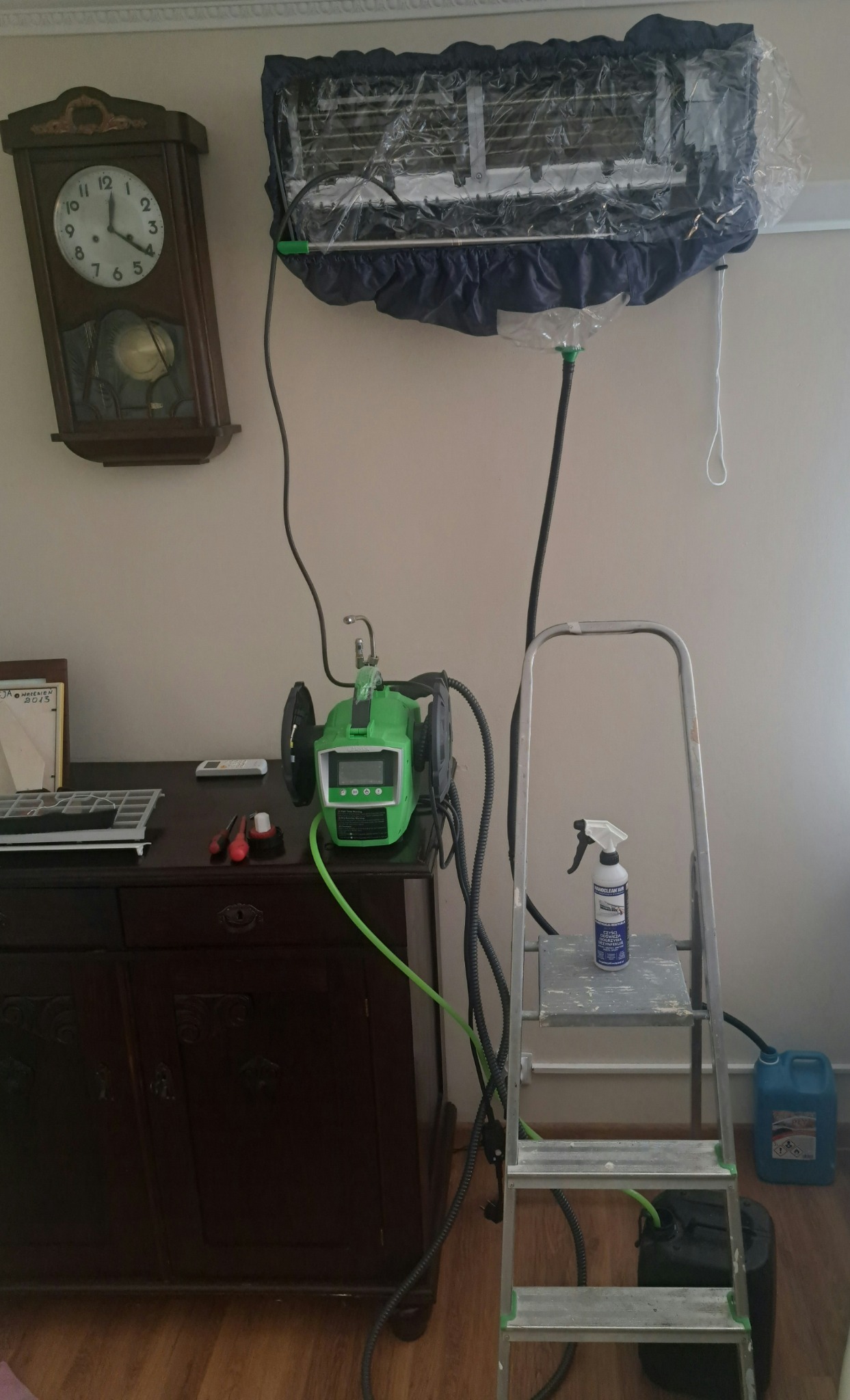 Czyszczenie klimatyzatora w domu: klimatyzator zabezpieczony folią ochronną z podpiętym urządzeniem do czyszczenia, drabina z butelką środka czyszczącego obok, na komodzie zegar i narzędzia.