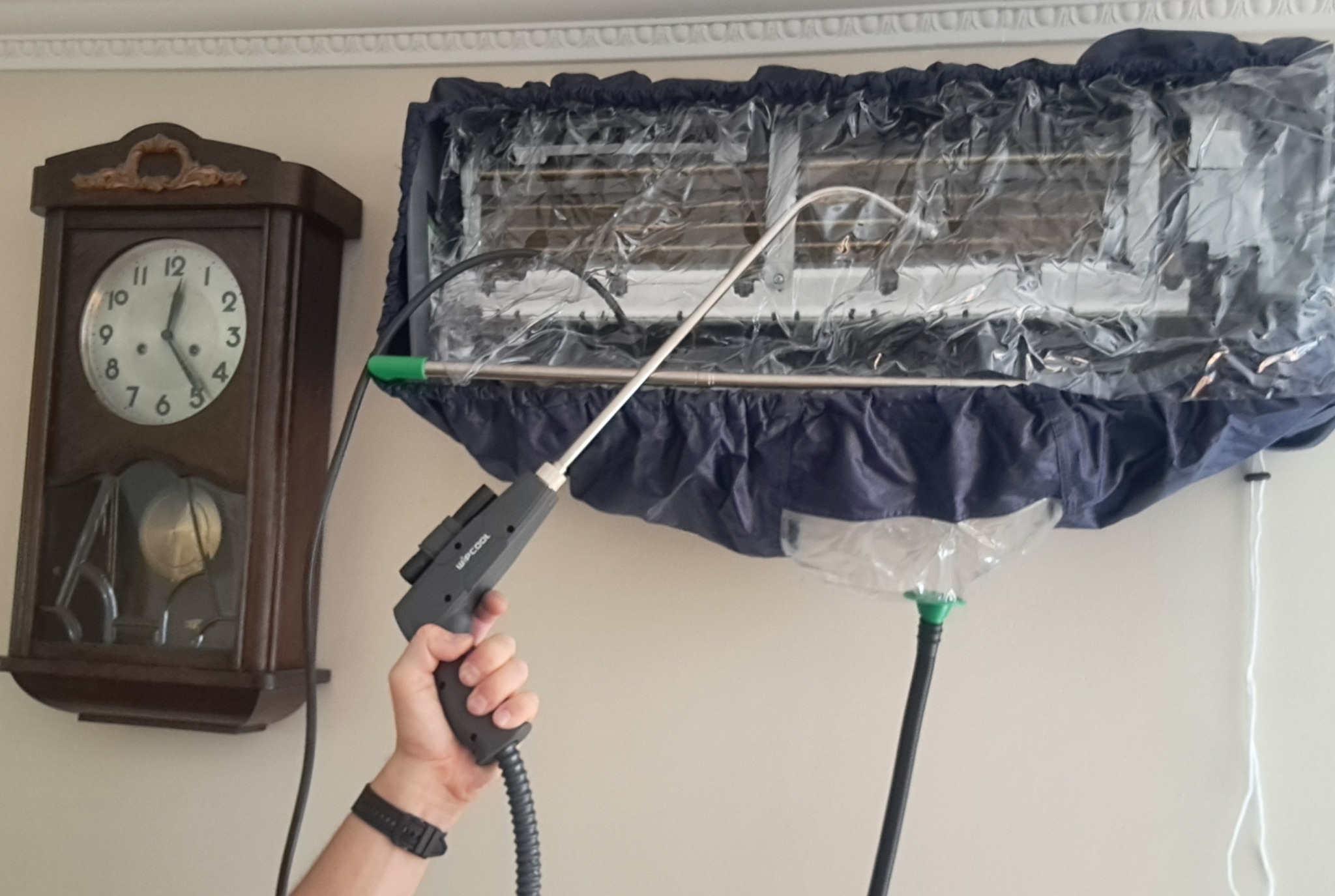 Czyszczenie klimatyzatora typu split za pomocą specjalistycznego urządzenia pod ciśnieniem, z założonym pokrowcem ochronnym zbierającym brud, obok wisi stary zegar.