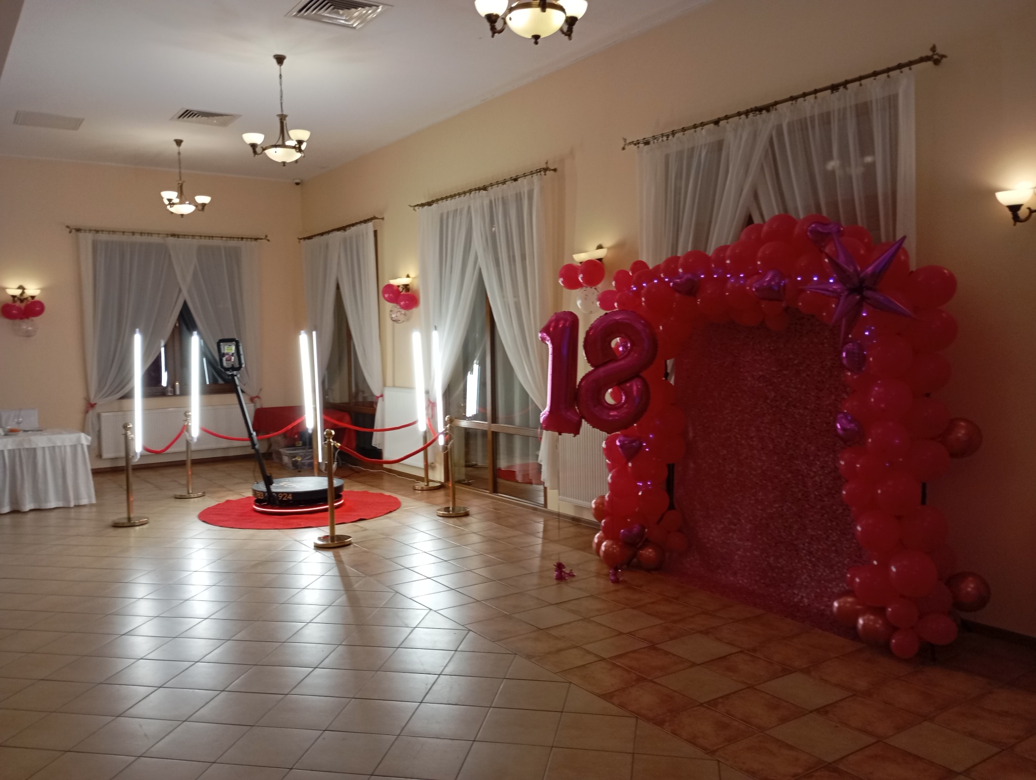 Sala bankietowa z dekoracjami urodzinowymi i obrotową platformą do nagrywania filmów 360 stopni, oświetlona lampami LED i otoczona słupkami z czerwonymi sznurami.