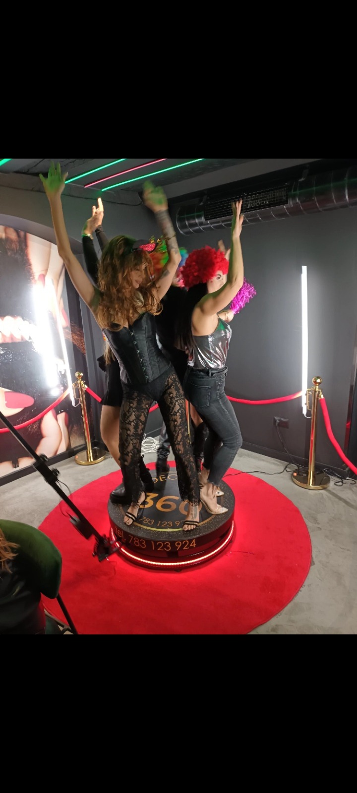 Trzy osoby w kostiumach i perukach pozują z podniesionymi rękami na obrotowej platformie z oświetleniem LED i numerem telefonu, otoczonej czerwonym dywanem i złotymi słupkami.