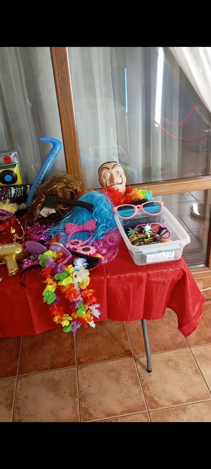 Różnorodne rekwizyty na stole z czerwonym obrusem: maska Dalego, kolorowe peruki, hawajskie naszyjniki, zabawkowy pistolet, okulary i inne akcesoria.