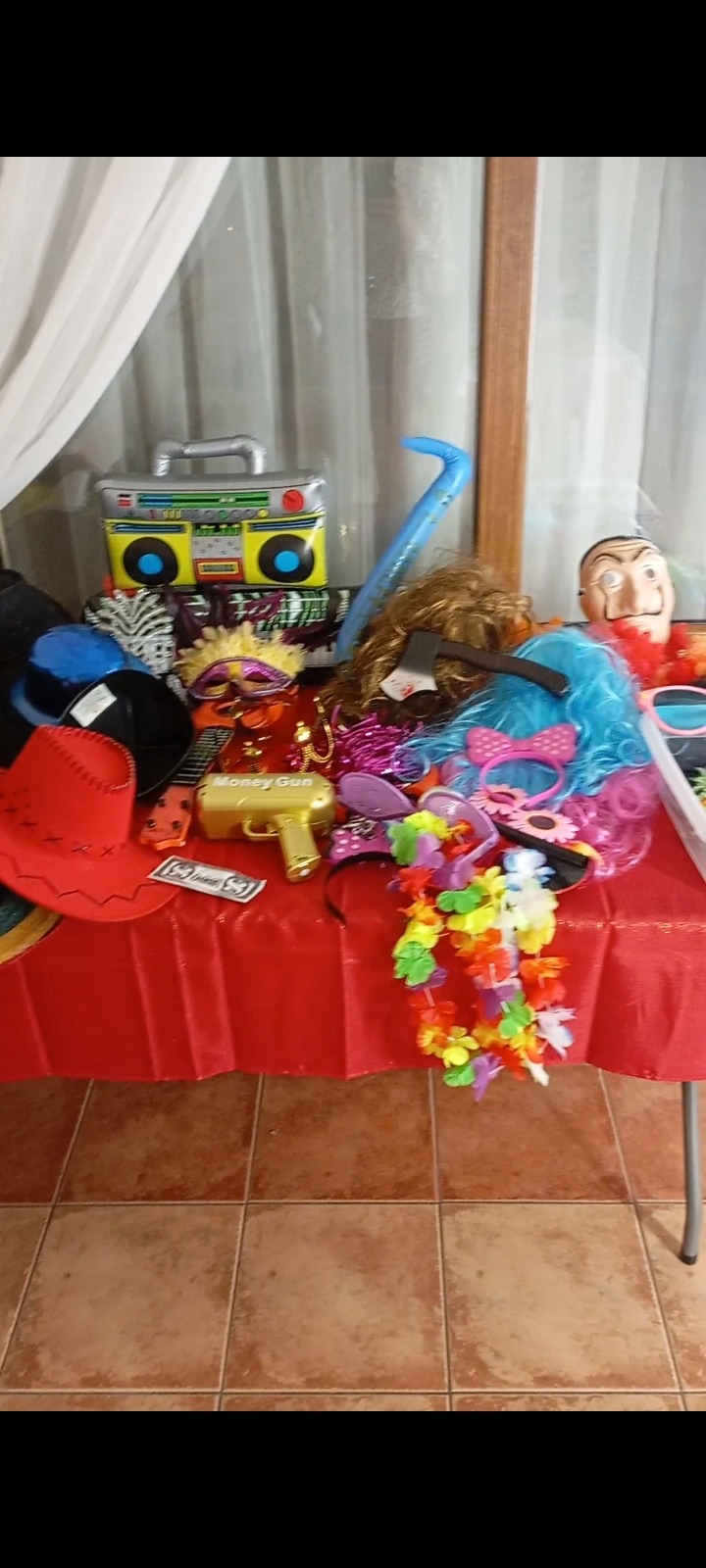 Różnorodne rekwizyty na stole z czerwonym obrusem: dmuchany boombox, kolorowe peruki, kapelusze, hawajski naszyjnik i zabawkowy pistolet na pieniądze.