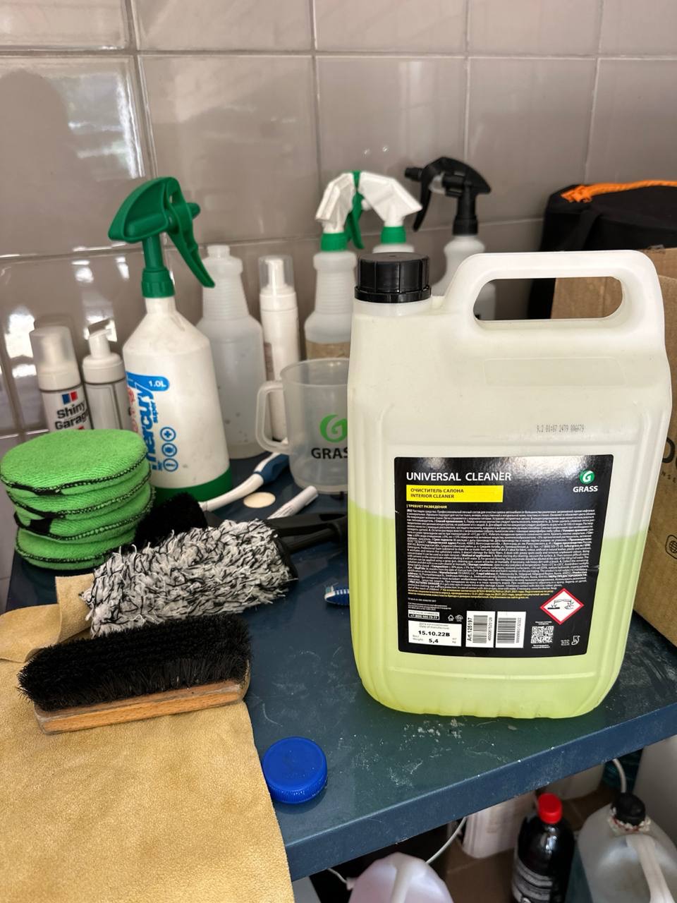 Kanister uniwersalnego środka czyszczącego Grass obok szczotek, ściereczek i spryskiwaczy na niebieskim stole w pomieszczeniu z kafelkami.