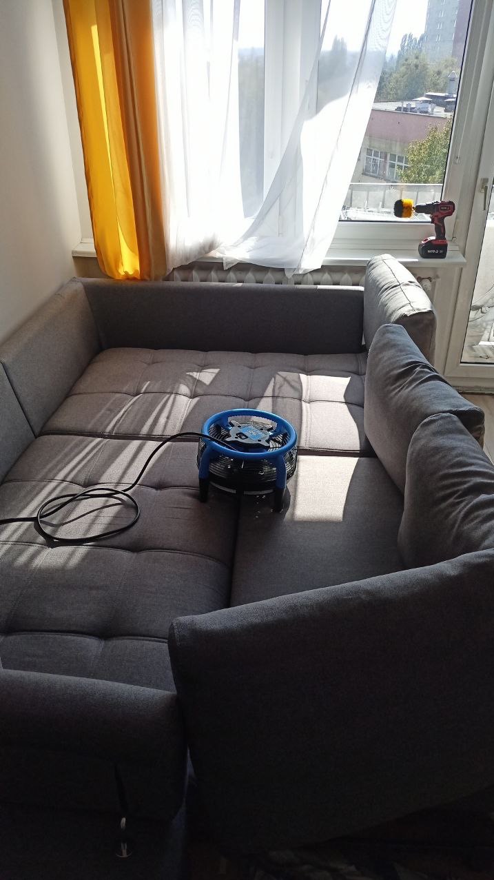 Szary, modułowy narożnik z niebieskim wentylatorem na siedzisku, obok okna z widoczną wiertarką na parapecie.