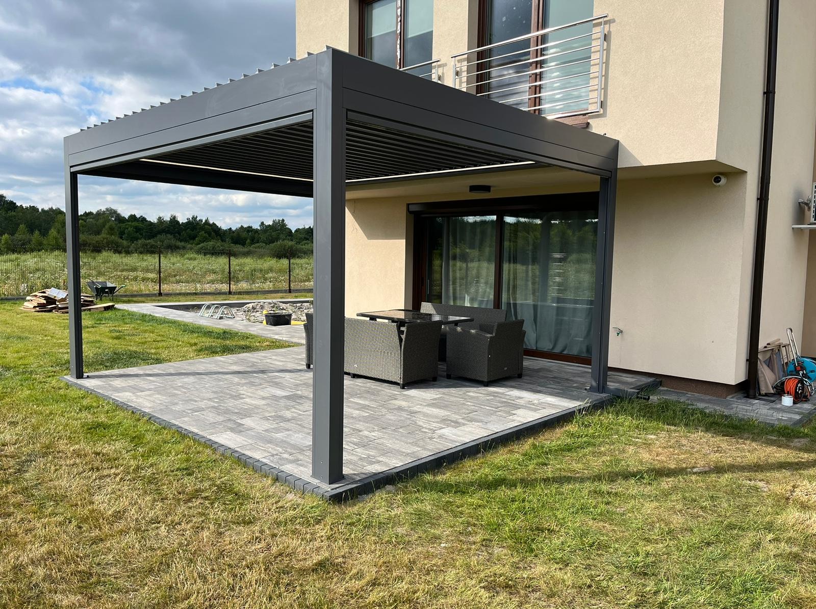 Nowoczesna, grafitowa pergola lamelowa przylegająca do domu z beżową elewacją, wyposażona w komplet mebli ogrodowych na szarym tarasie z kostki brukowej. W tle widoczny trawnik i zieleń drzew.