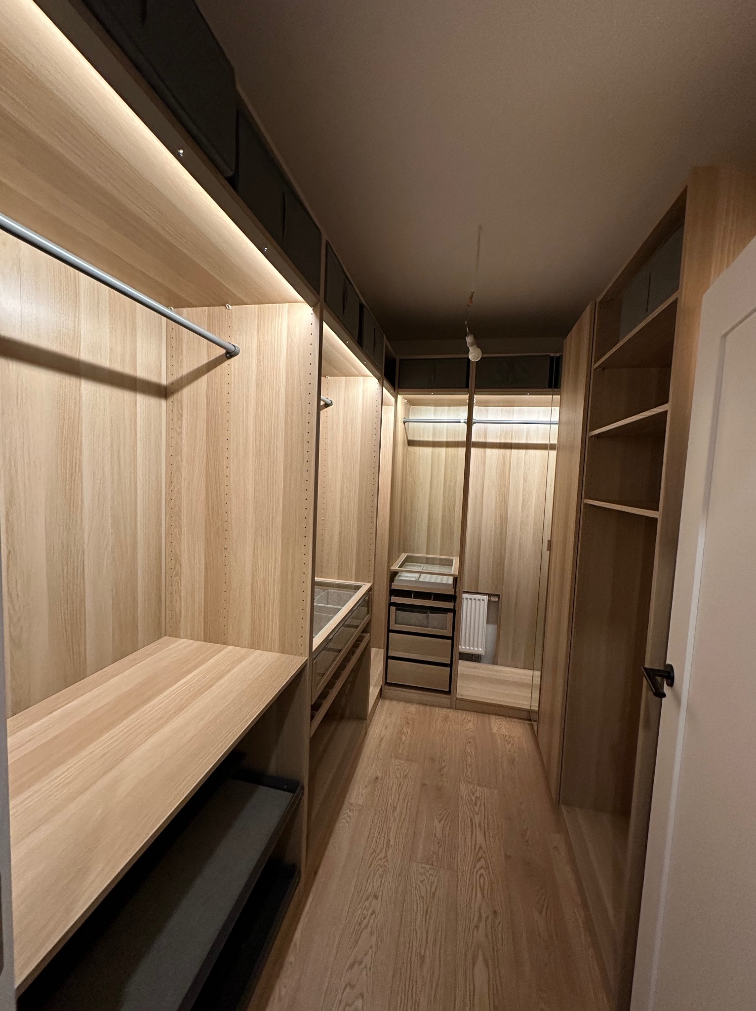 Wnętrze przestronnej garderoby z jasnego drewna, z półkami, szufladami i drążkami na ubrania, oświetlenie LED w górnej części, widoczny fragment podłogi z paneli w odcieniu drewna.