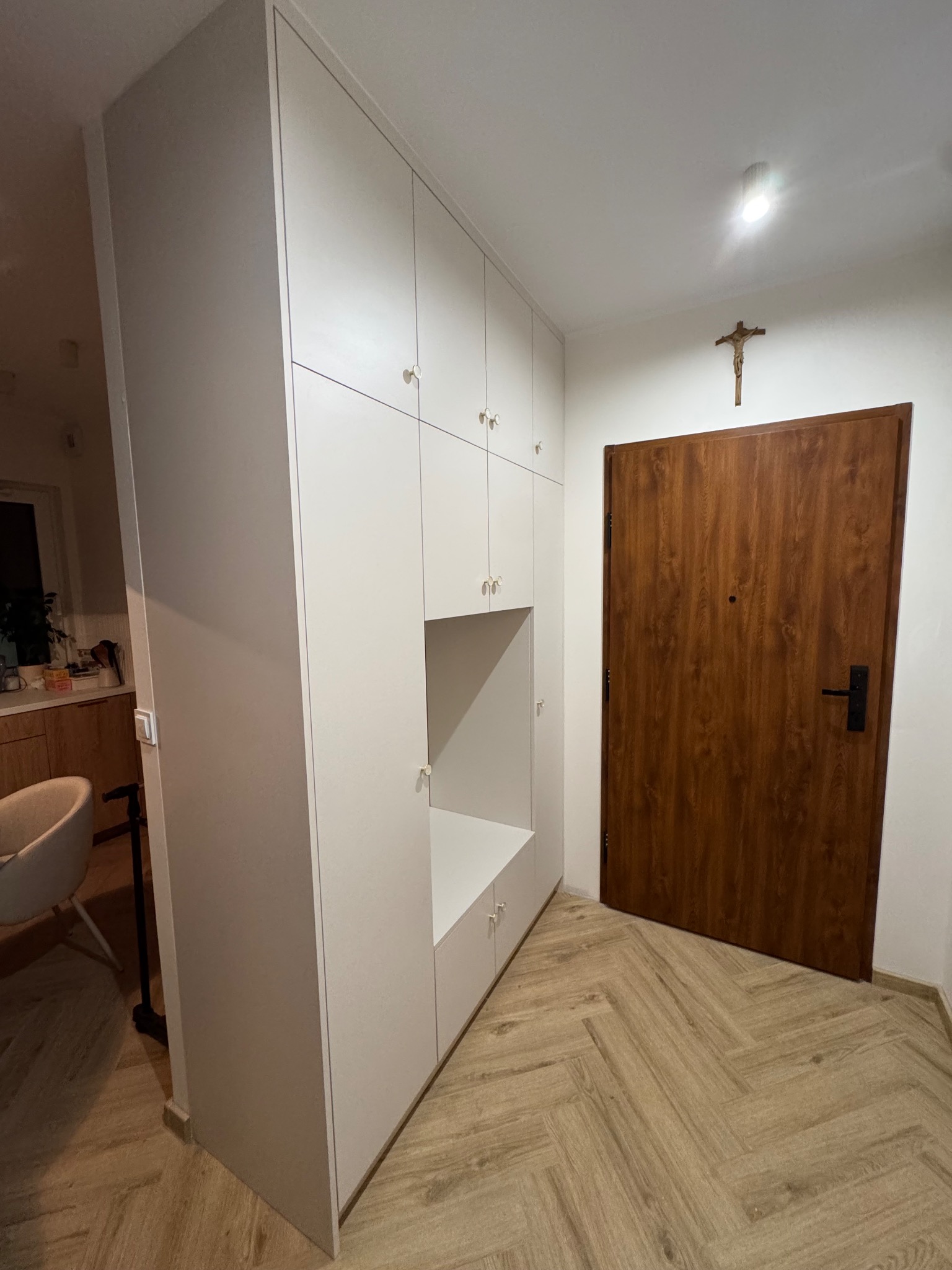 Biała szafa wnękowa z otwartą półką i złotymi uchwytami, dopasowana do przedpokoju z drewnianą podłogą w jodełkę i brązowymi drzwiami.