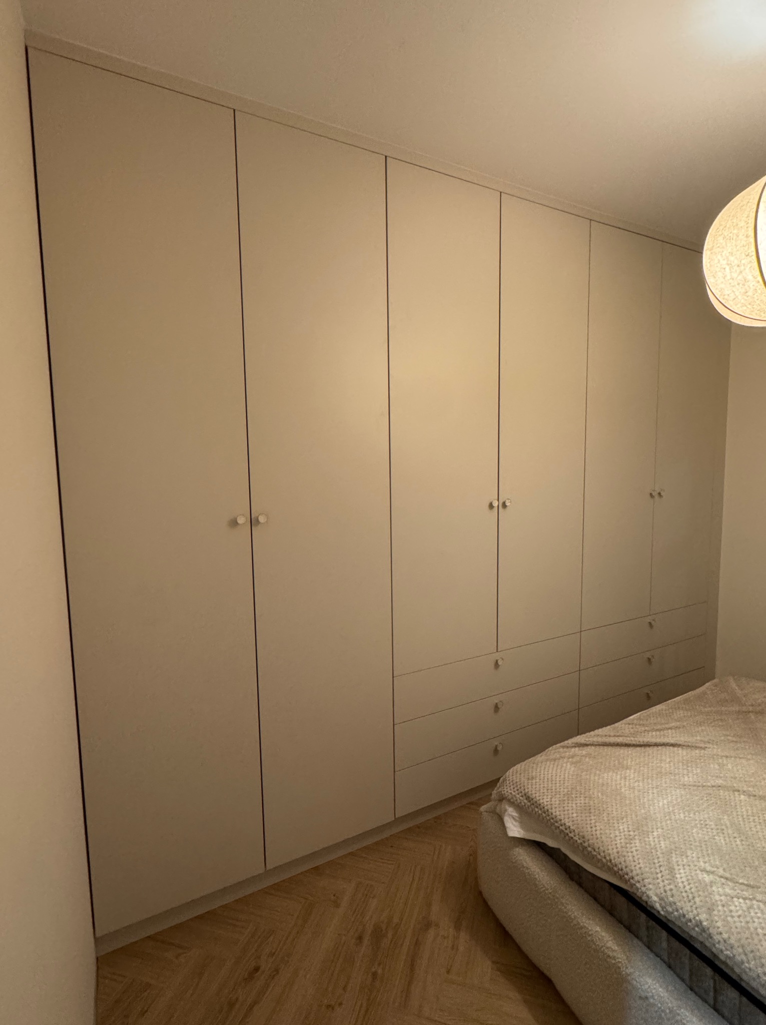 Jasna, zabudowana szafa z szufladami i okrągłymi uchwytami, dopasowana do wnętrza sypialni z parkietem i widocznym fragmentem łóżka.