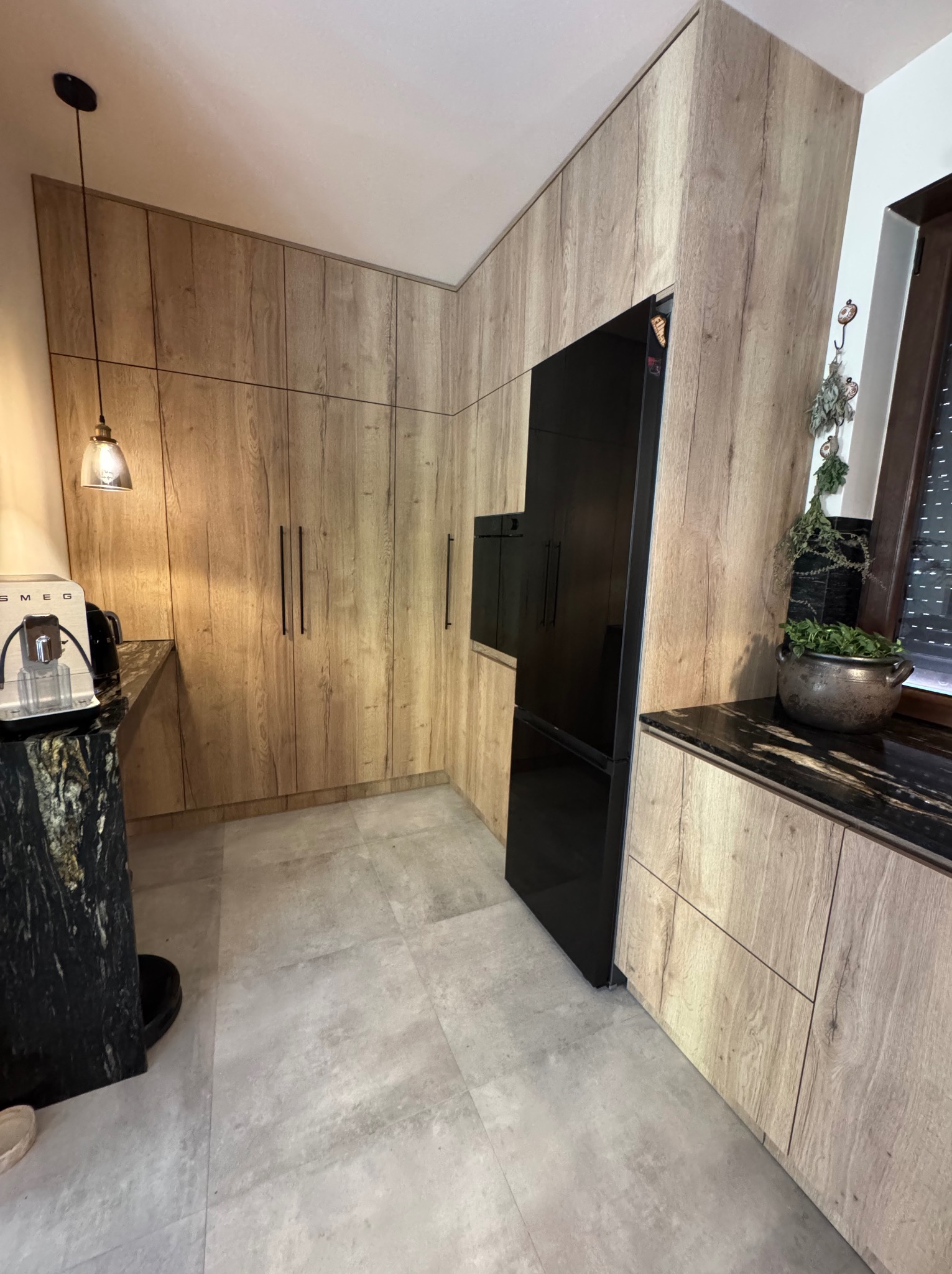 Kuchnia na wymiar w Poznaniu: zabudowa z drewnopodobnymi frontami, czarny sprzęt AGD i blat z ciemnego kamienia. Nowoczesny design z wiszącą lampą.