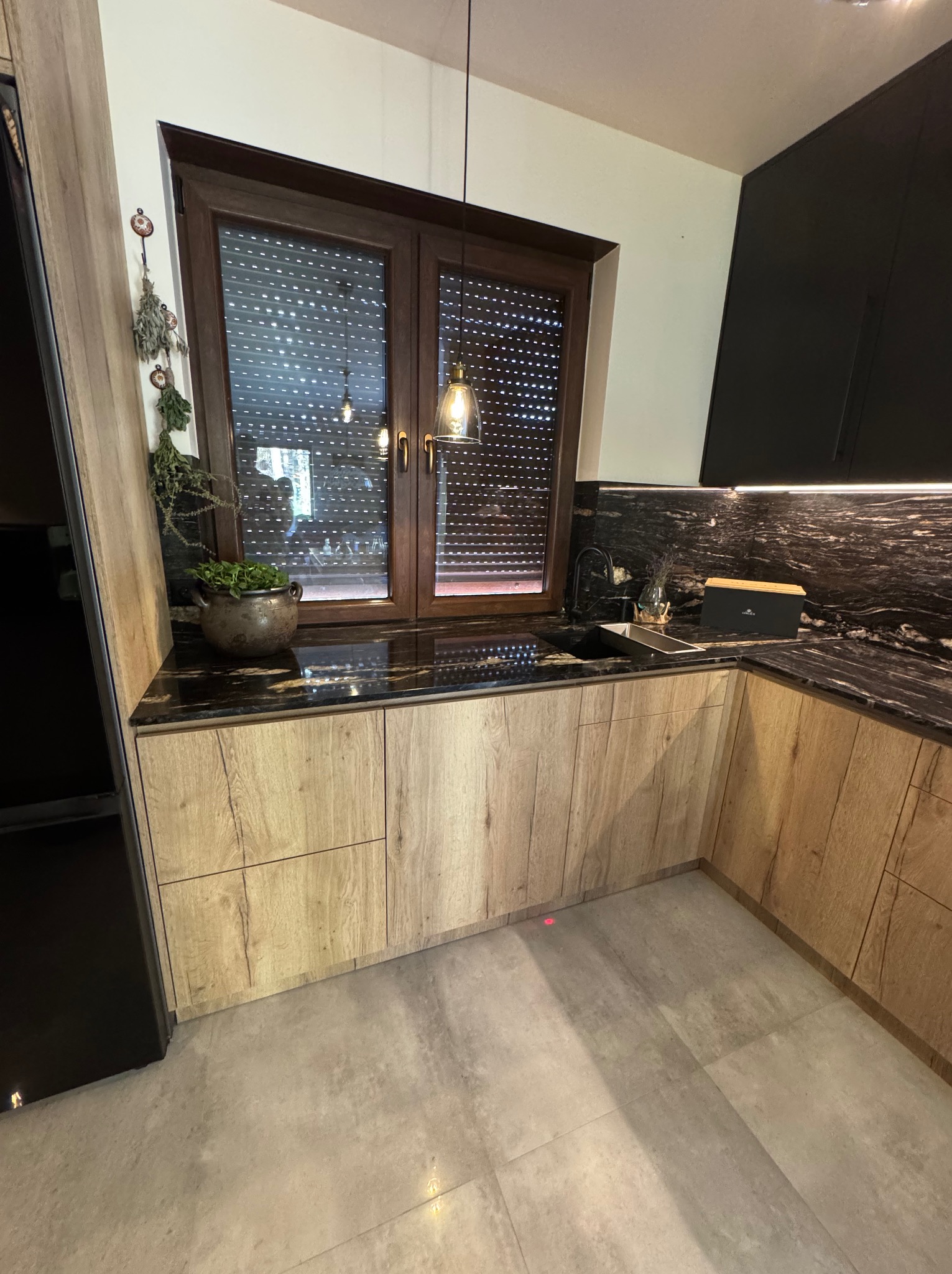 Nowoczesna kuchnia z drewnianymi frontami szafek, czarnym blatem z marmurkowym wzorem i oknem z żaluzjami. Uwagę przykuwa doniczka z ziołami na blacie.