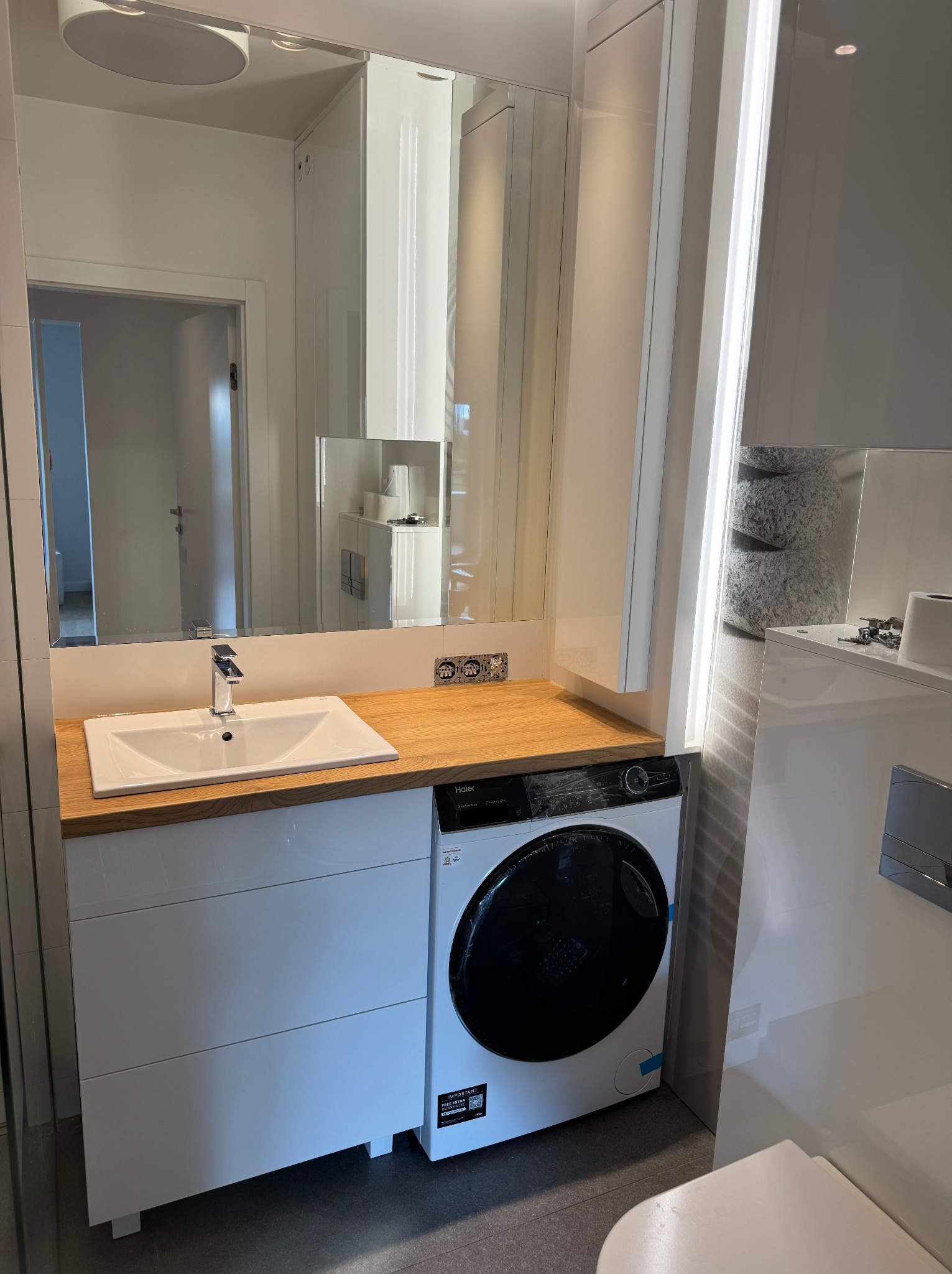 Nowoczesna łazienka z szafką pod umywalkę z białymi frontami i drewnianym blatem. Obok pralka w zabudowie. Lustro z oświetleniem LED. Minimalistyczny design.