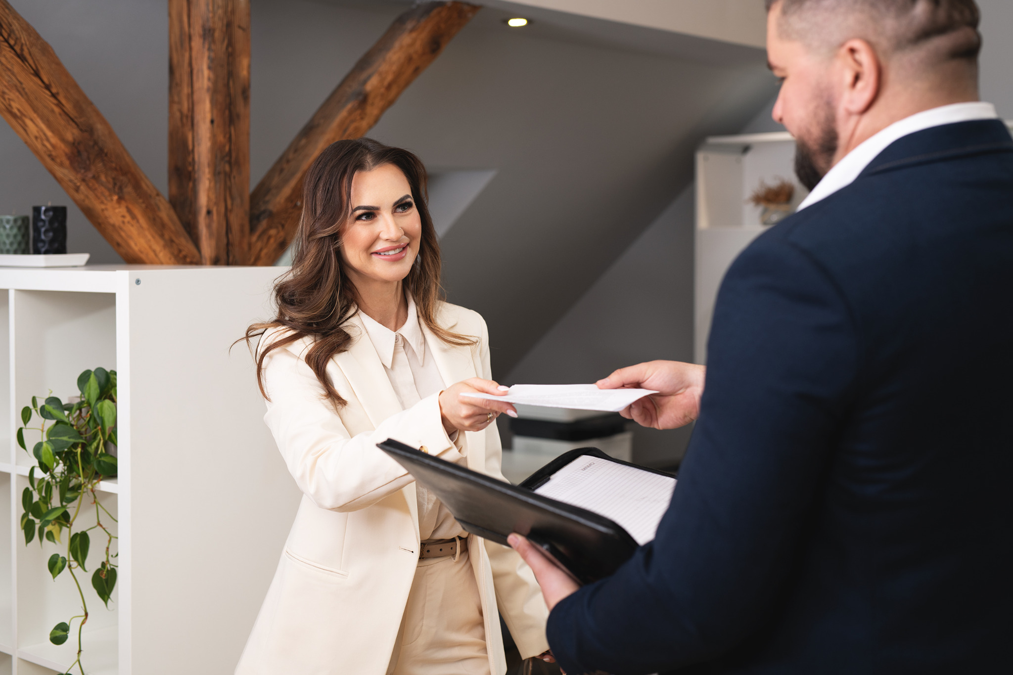 Kobieta w kremowym garniturze przekazuje dokument mężczyźnie w granatowym garniturze, który trzyma otwarty segregator z notatkami.