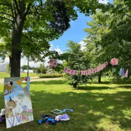 Dekoracje urodzinowe w plenerze: girlanda 'Happy Birthday' zawieszona między drzewami, fotobudka z motywami zwierzęcymi i rekwizyty do zdjęć rozłożone na trawie w słoneczny dzień.
