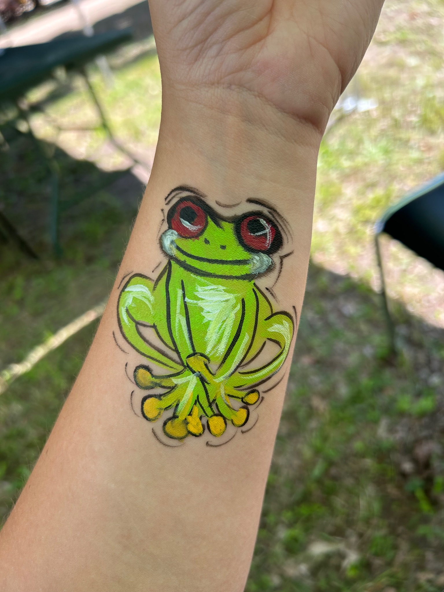 Malunek na ramieniu przedstawiający zieloną żabę z czerwonymi oczami i żółtymi palcami, wykonany farbami do ciała.