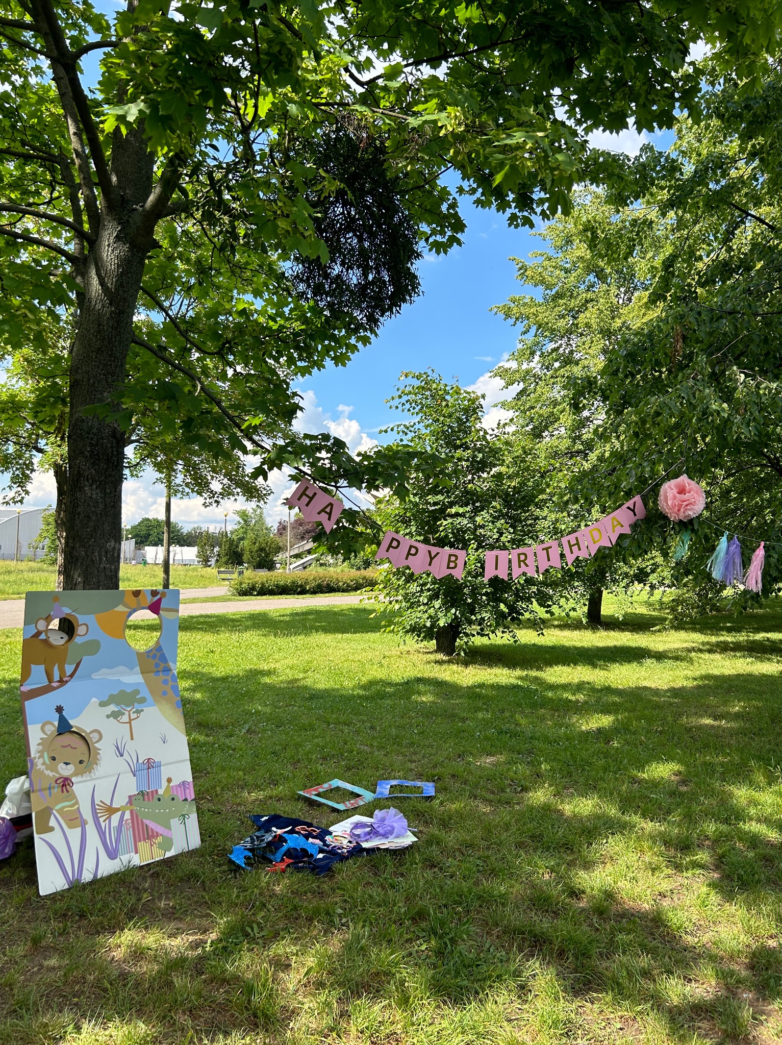 Dekoracje urodzinowe w plenerze: girlanda 'Happy Birthday' zawieszona między drzewami, fotobudka z motywami zwierzęcymi i rekwizyty do zdjęć rozłożone na trawie w słoneczny dzień.