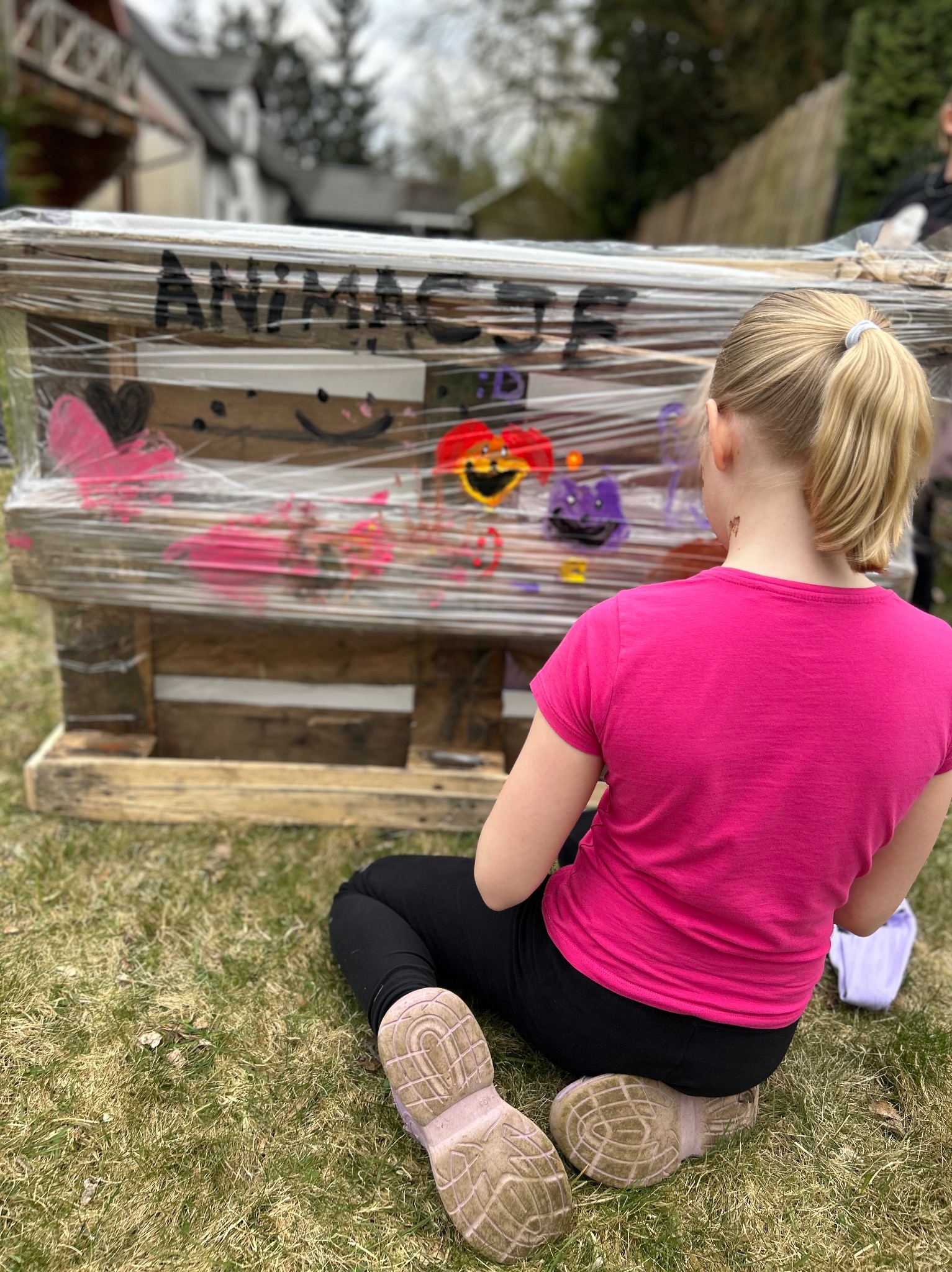 Dziewczynka w różowej koszulce maluje kolorowe postacie na drewnianej konstrukcji z napisem 'ANIMACJE', owiniętej folią, siedząc na trawie. Kreatywne zajęcie.