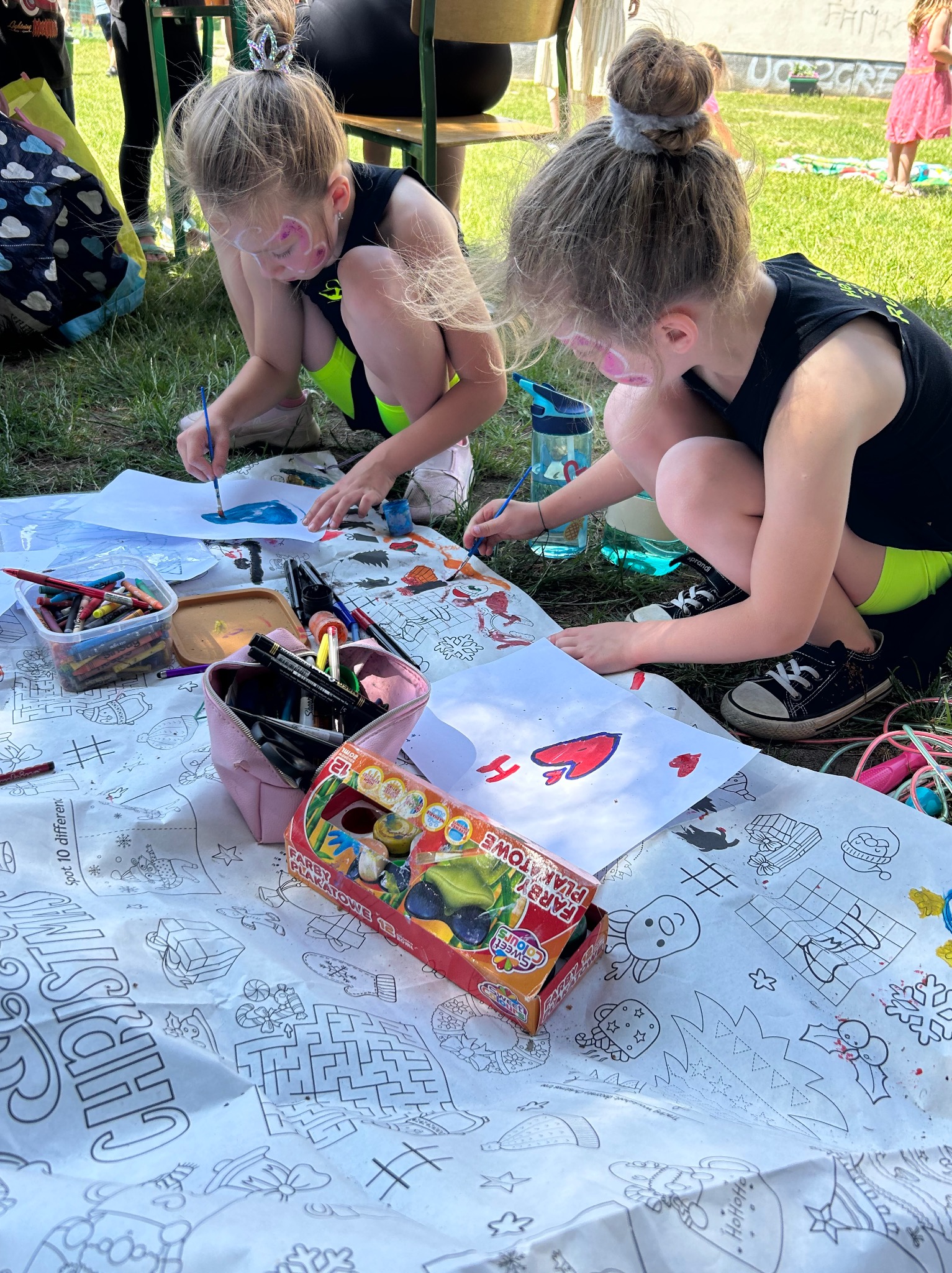 Dwie dziewczynki z pomalowanymi twarzami, siedząc na trawie, kolorują świąteczne kolorowanki farbami plakatowymi. Widoczne kredki, pędzle i pudełko z farbami.