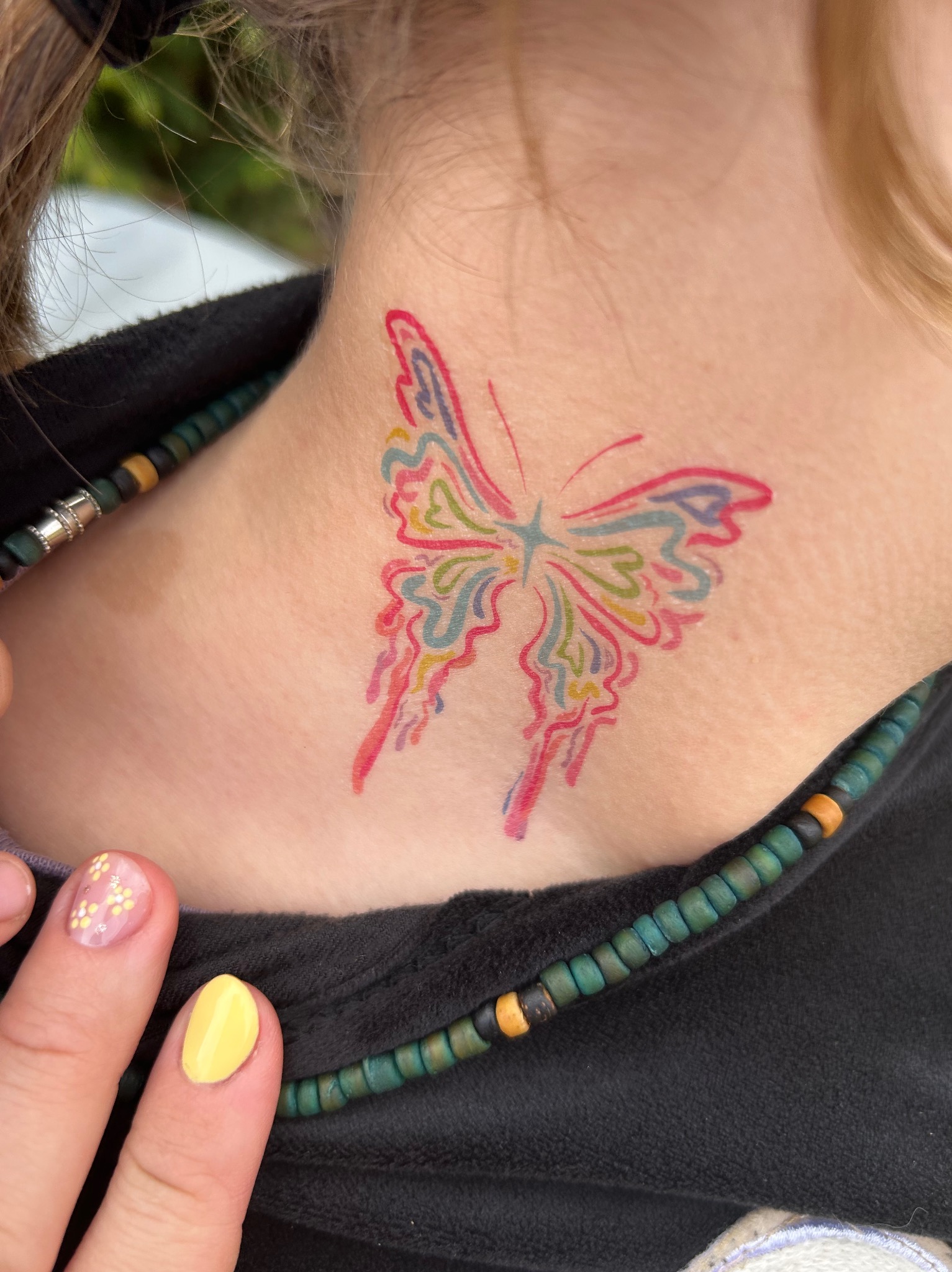 Kolorowy tatuaż motyla na szyi dziewczyny, widoczny fragment naszyjnika z koralików i dłoni z pomalowanymi paznokciami. Zbliżenie na detale.
