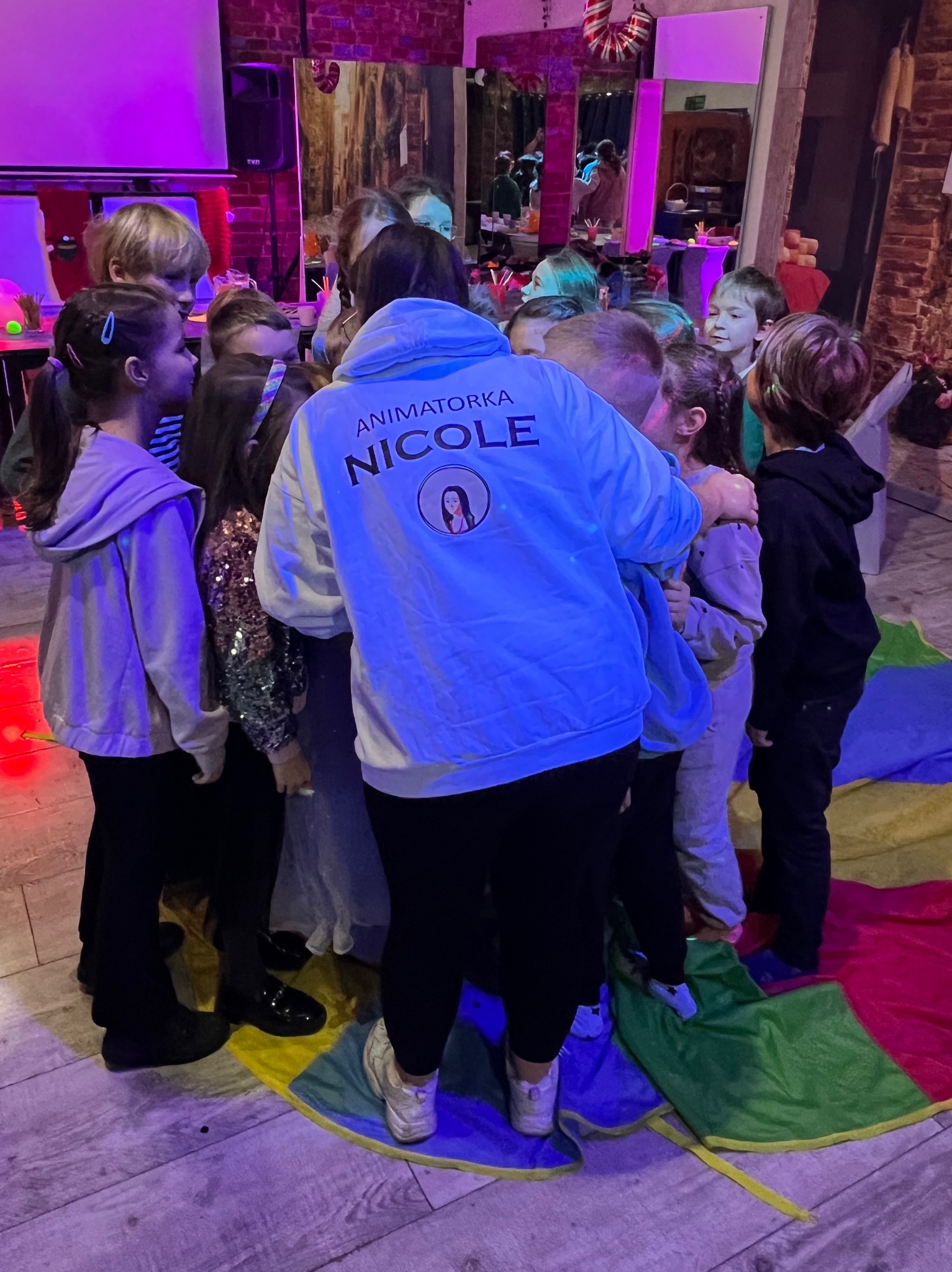 Grupka dzieci otoczona przez animatora w bluzie z napisem 'Animator NICOLE' podczas zabawy z kolorową chustą animacyjną w pomieszczeniu z ceglaną ścianą.