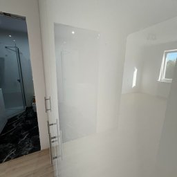 FIX WOOD ZABUDOWY MEBLOWE - Nowoczesna łazienka z czarnym marmurem i kabiną prysznicową za szklanymi drzwiami z metalowymi uchwytami, widok na jasne, puste pomieszczenie z oknem.