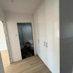 FIX WOOD ZABUDOWY MEBLOWE - Biała szafa wnękowa z połyskiem w nowym mieszkaniu, widok na marmurową łazienkę z kabiną prysznicową i wanną, podłoga wyłożona panelami.