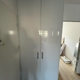 FIX WOOD ZABUDOWY MEBLOWE - Wysoka, biała szafa wnękowa z połyskiem, z minimalistycznymi uchwytami, zamontowana w pokoju z panelami podłogowymi; widoczne elementy wykończeniowe w tle.