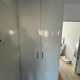 Wysoka, biała szafa wnękowa z połyskiem, z minimalistycznymi uchwytami, zamontowana w pokoju z panelami podłogowymi; widoczne elementy wykończeniowe w tle.